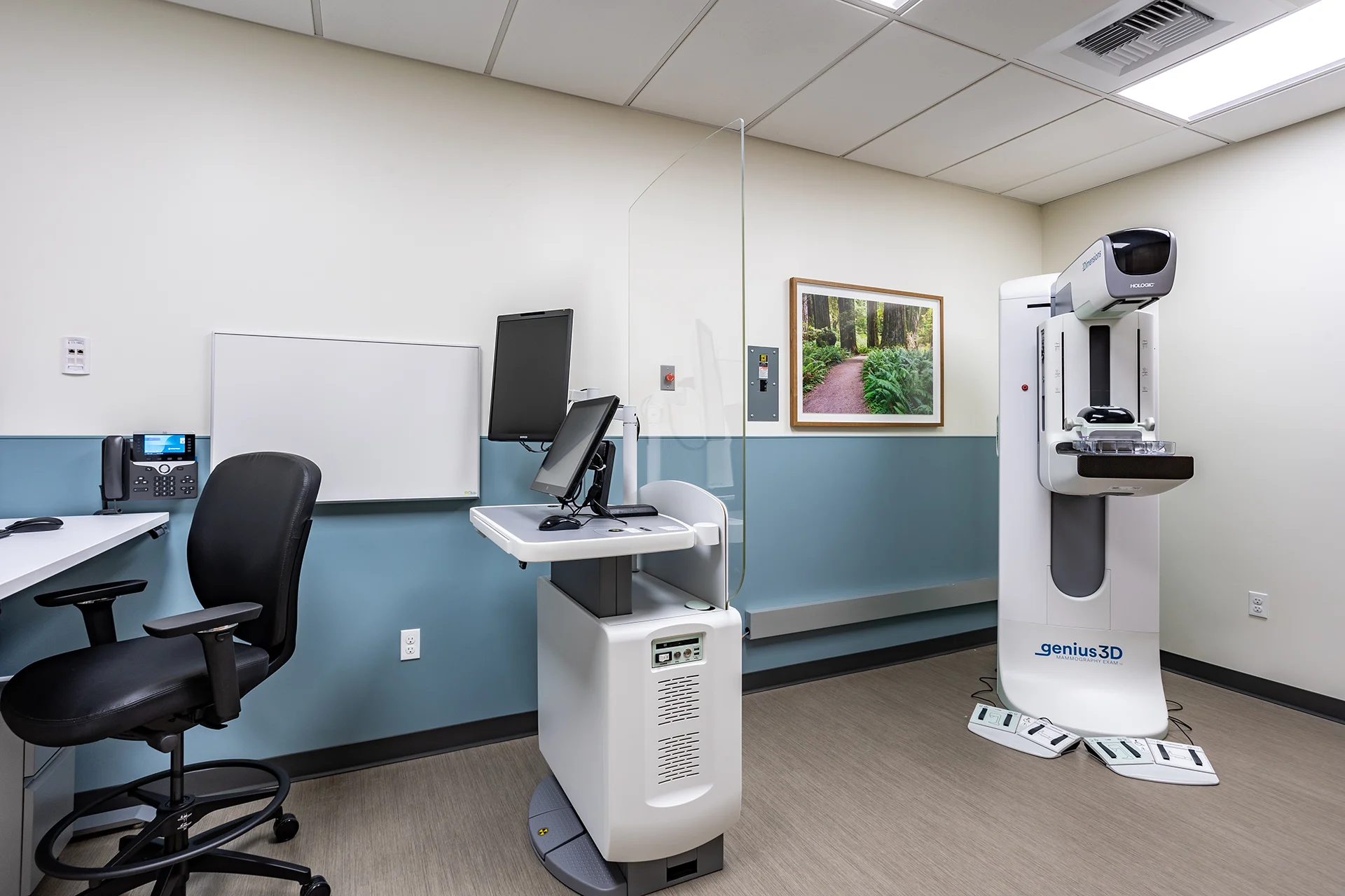 Advanced breast imaging suite featuring a Hologic Genius 3D system, a mobile control console behind an integrated clear radiation protection barrier, a wall-mounted porcelain whiteboard, blue accent wainscoting, and institutional resilient flooring.