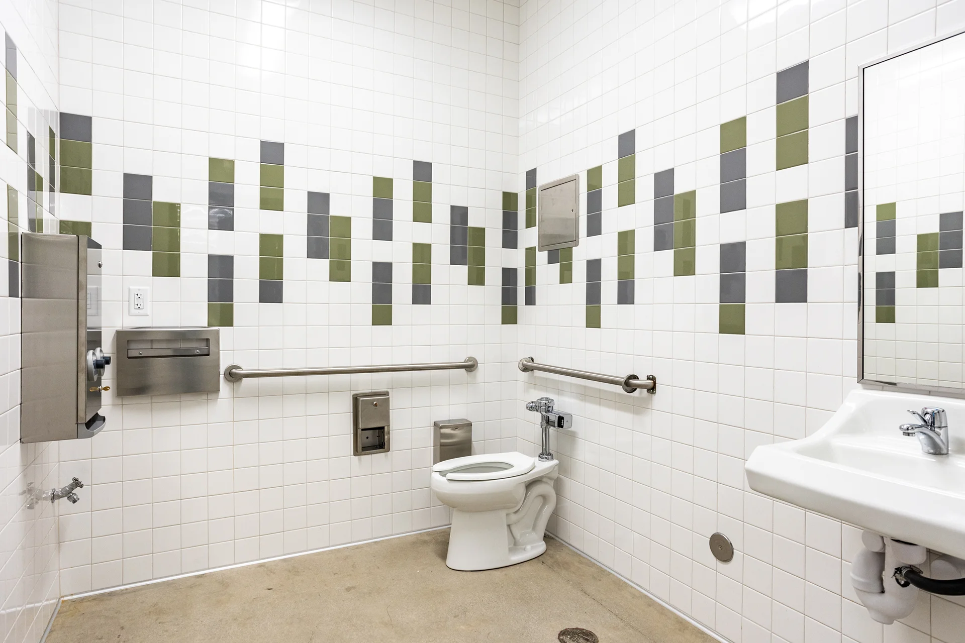 Interior view of a fully accessible institutional restroom, showcasing a white porcelain toilet fixture with dual stainless steel grab bars, a wall-hung sink basin, concrete flooring, and decorative vertical bands of gray and green ceramic subway tiles.