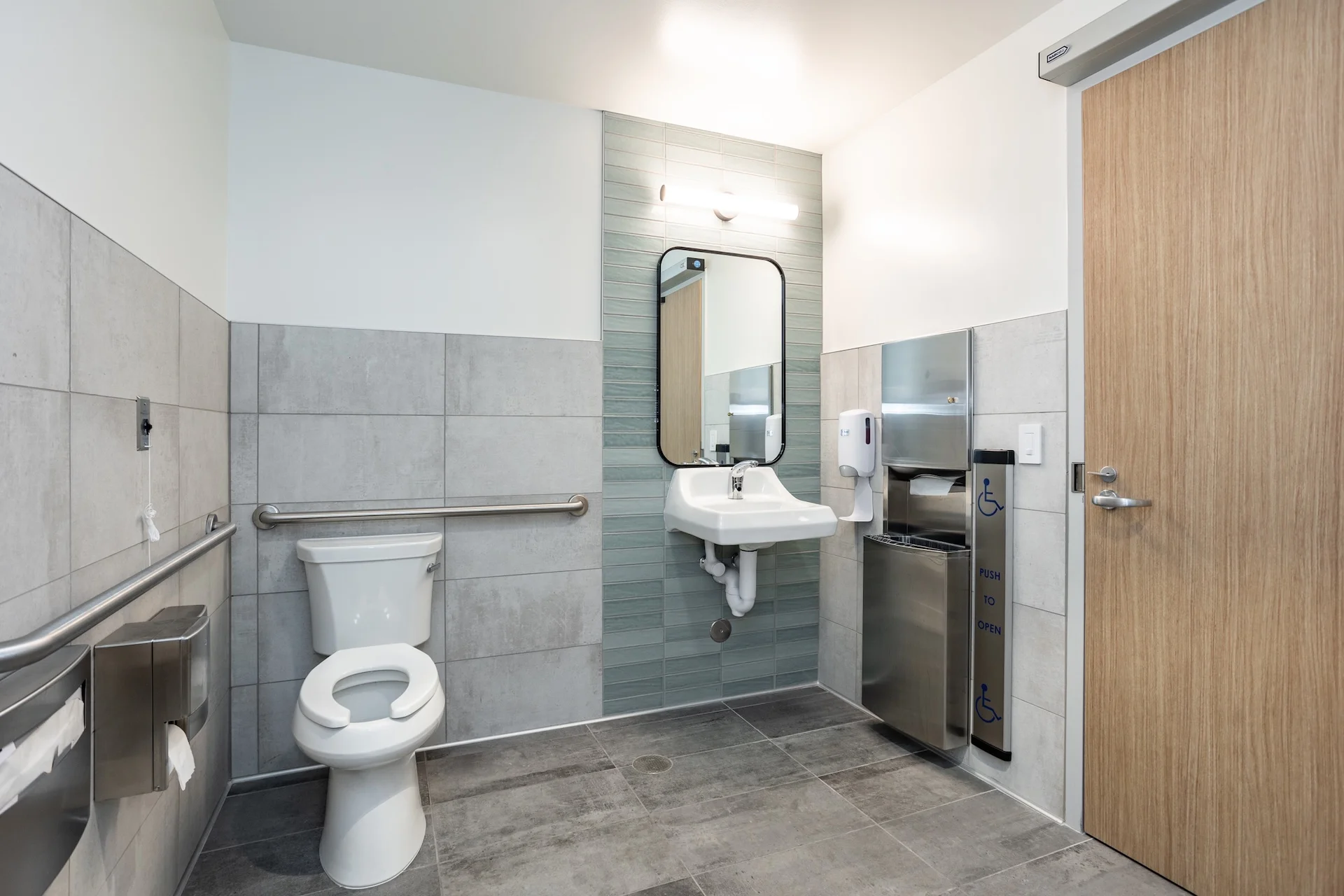Accessible medical clinic restroom showcasing a wall-mounted porcelain sink, a vertical stack of seafoam green glass subway tile, stainless steel grab bars, a commercial flush-valve toilet, large-format gray porcelain floor tiles, and a wide-clearance automatic sliding wood door.