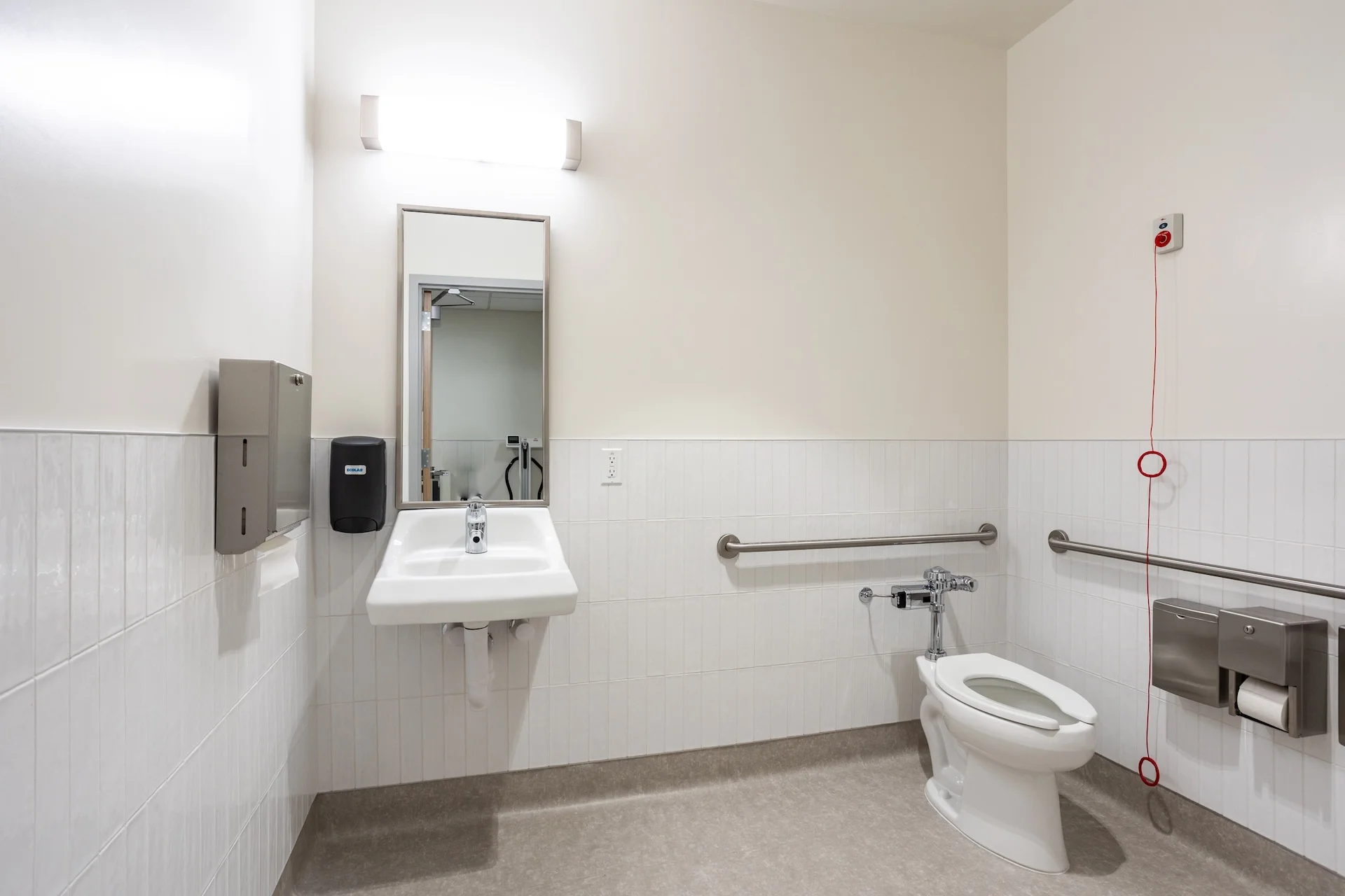 Interior view of a barrier-free commercial bathroom within a medical center, featuring a wall-hung sink, an ADA-height toilet with stainless steel grab bars, a smooth-surface resilient floor, white tiled wainscoting, and an emergency pull cord system.
