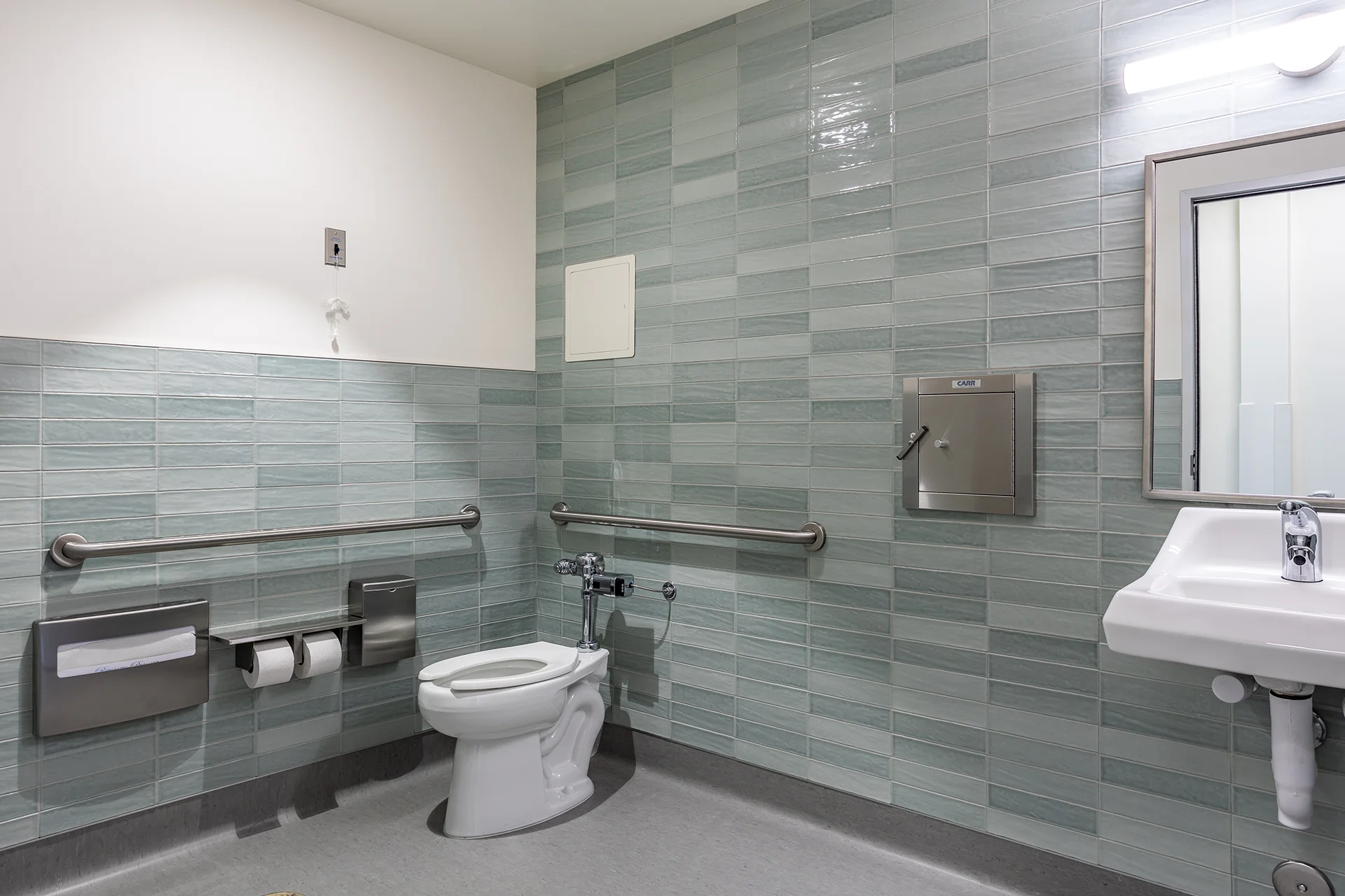 Institutional commercial bathroom design displaying a floor-mounted elongated toilet with an automatic flush valve, wrap-around stainless steel grab bars, a vitreous china basin sink, a stainless steel waste receptacle, and full-height seafoam green glass subway tile wainscoting.