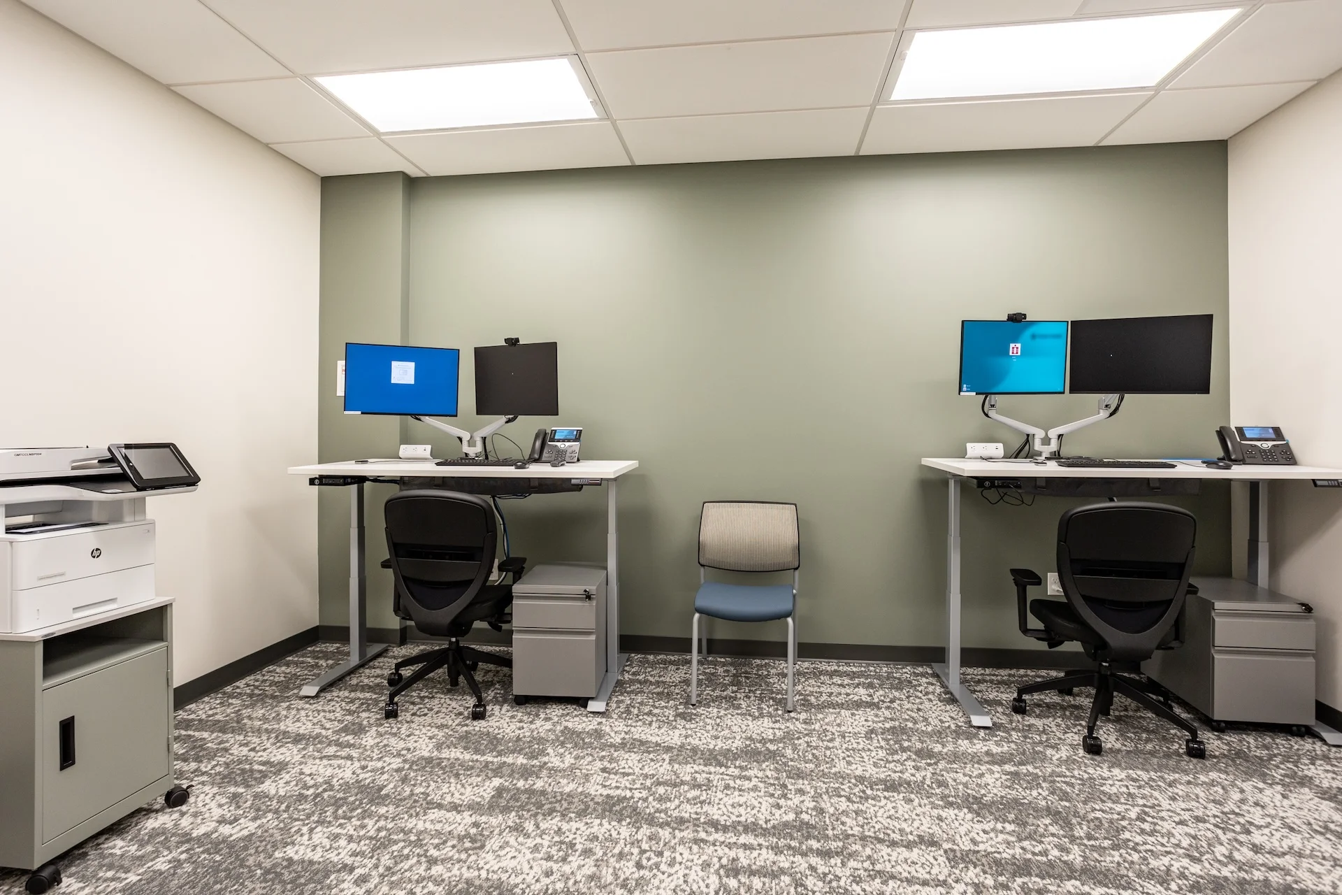 Commercial healthcare office space featuring dual height-adjustable sitting-to-standing desks with ergonomic black rolling chairs, mounted computer monitors, dedicated business telephones, under-desk file cabinets, textured commercial-grade carpeting, and a sage green accent wall.