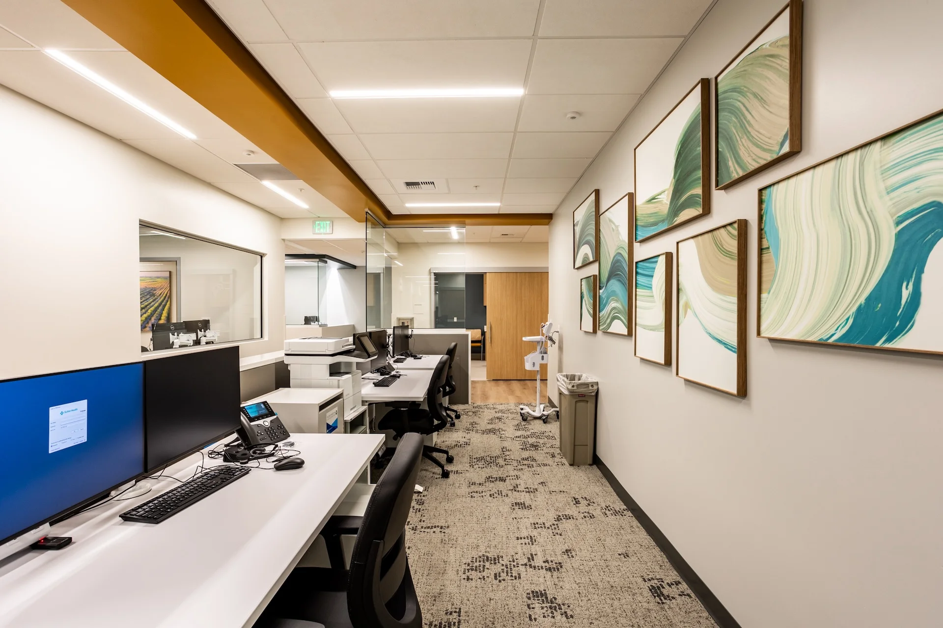 Back-office administrative area showing multiple networked workstations with flat-screen monitors, printer stations, an architectural ceiling soffit, patterned commercial carpet flooring, and a gallery-style wall displaying framed canvas artwork.