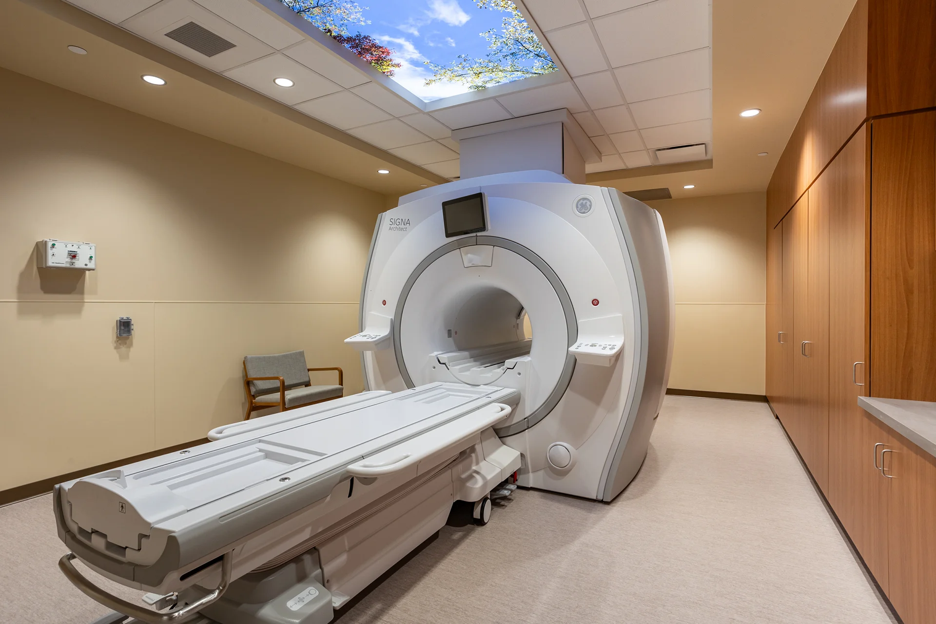 Comprehensive view of a modern MRI examination room highlighting a large central scanner machine, a custom illuminated virtual sky ceiling panel, built-in natural wood laminate storage closets, and beige vinyl composition tile flooring.
