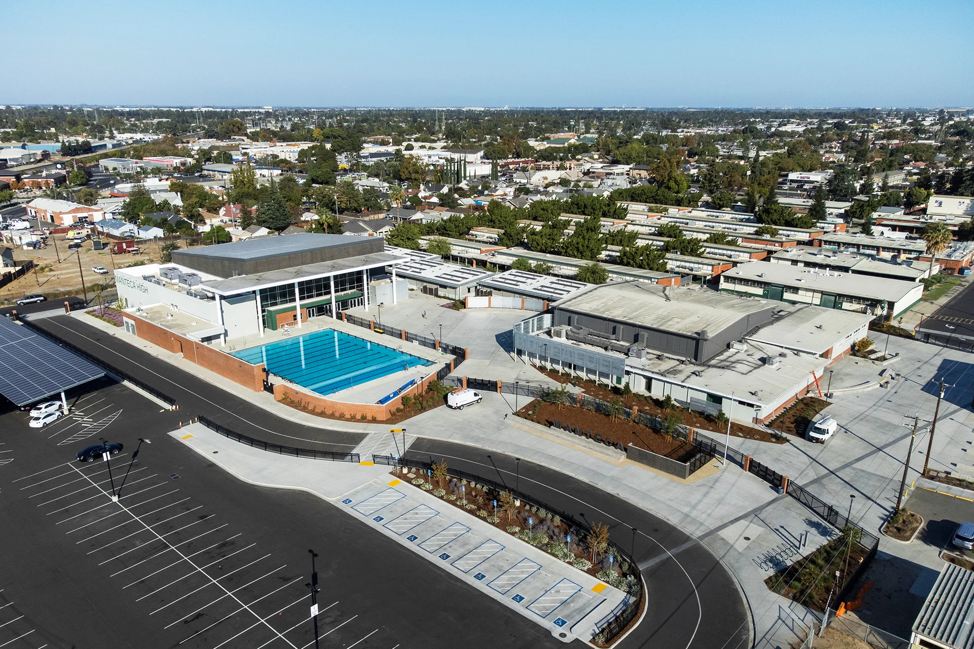 Wide-angle high-altitude drone perspective looking down at an institutional aquatics center facility in Manteca, California, showcasing an outdoor competitive swimming pool, flat roof surfaces with commercial mechanical HVAC infrastructure units, a newly asphalted vehicle parking field with designated ADA-compliant stalls, and clean concrete entry promenades extending across the campus footprint.