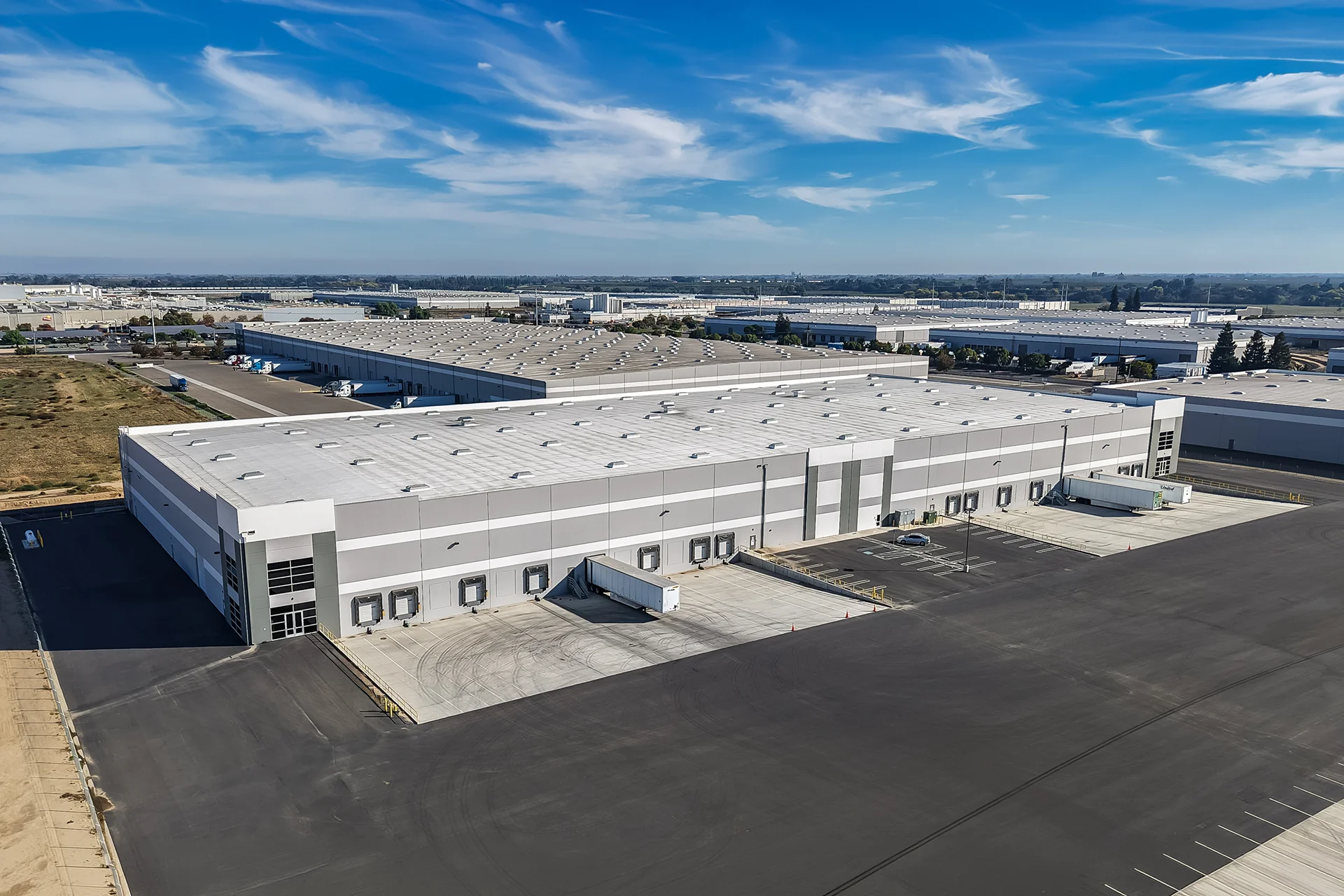 High-angle drone shot of a modern tilt-up concrete industrial distribution center, showcasing a low-slope white thermoplastic polyolefin membrane roof with rows of regular skylights, multiple recessed commercial loading docks with mechanical dock levelers, and an expansive asphalt logistics yard under a clear sky.