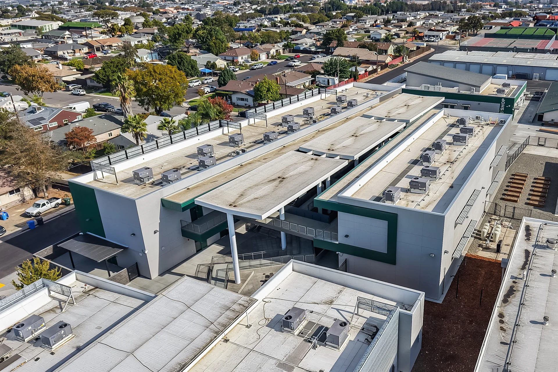 Overhead drone view of a newly developed commercial school facility in Manteca, California, showcasing white thermoplastic polyolefin TPO roofing sections lined with rows of packaged heat pump air conditioner condensers, structural steel courtyard connections, and surrounding residential neighborhoods under a clear afternoon sky.