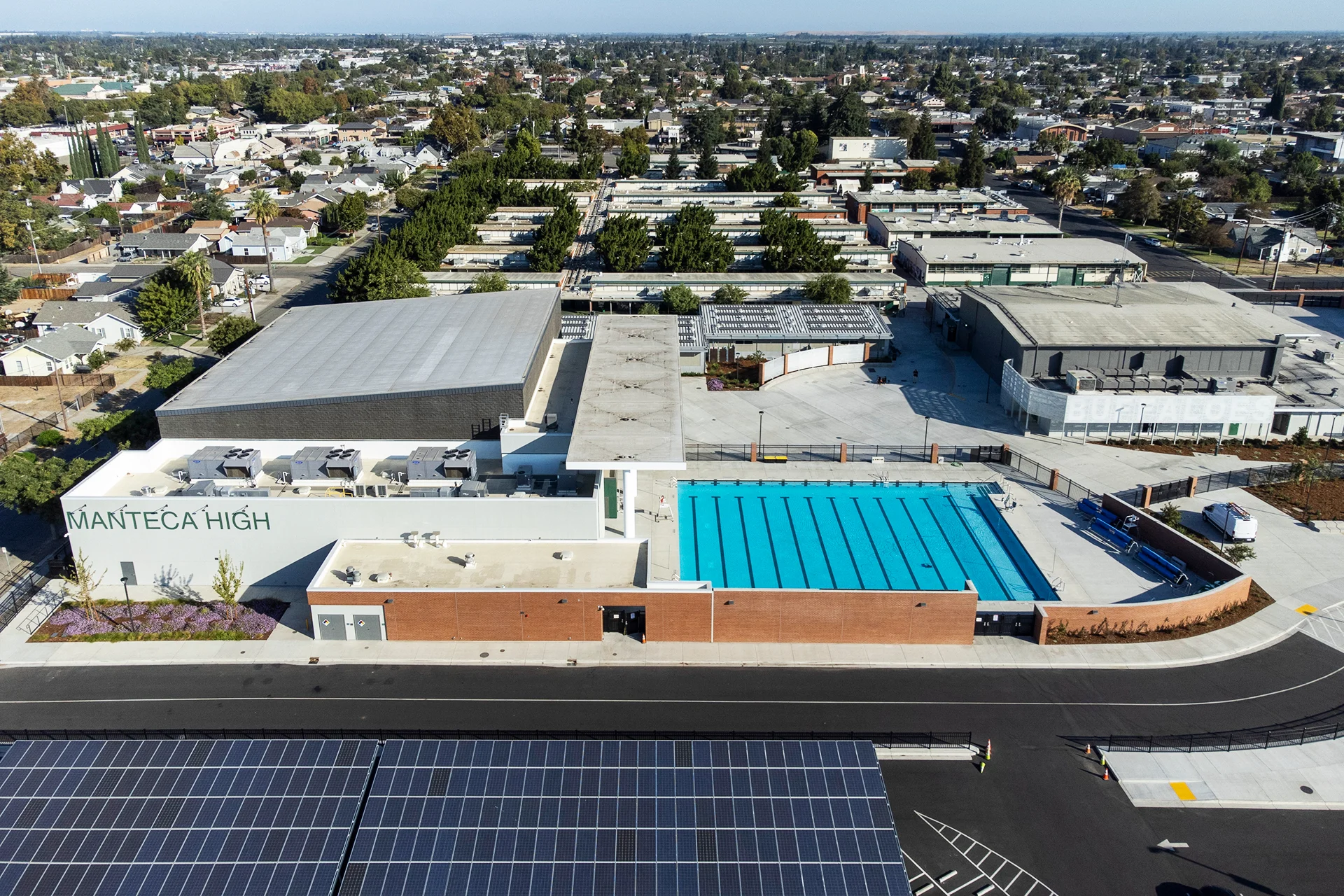 High-angle drone perspective looking down at a modern competitive swimming pool and gymnasium complex, highlighting flat roof membranes supporting commercial package unit HVAC systems, adjacent carports fitted with photovoltaic solar panels, and surrounding urban neighborhoods.