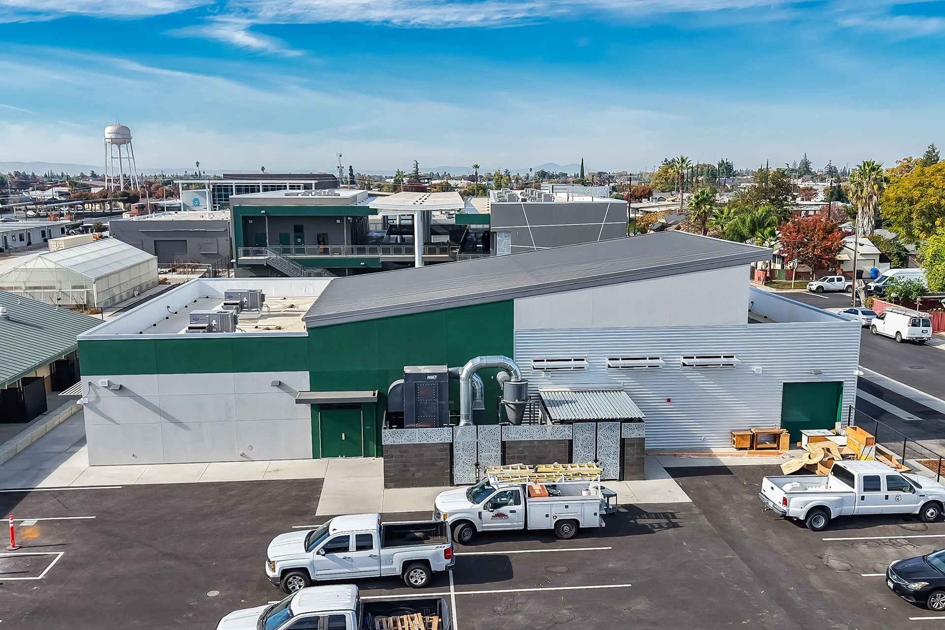 Expansive high-angle shot looking down at an institutional shop facility building, showcasing dark green and light grey stucco surfaces, a prominent exterior ventilation exhaust system, commercial work trucks parked in striped asphalt stalls, and a municipal water tower structure on the horizon.