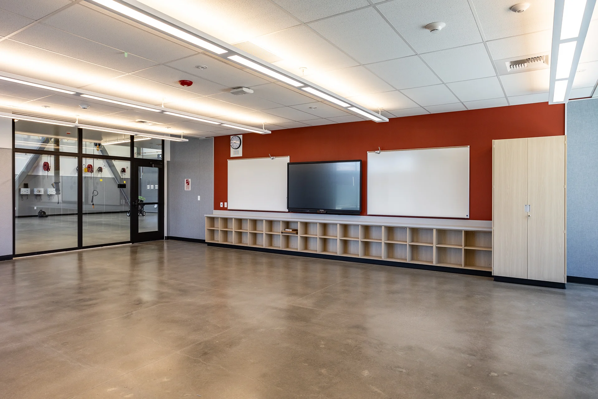 Wide-angle corner perspective of a modern educational workshop showing a red accent presentation wall with integrated open cubby storage, commercial glass partitioning walls revealing an adjacent mechanical bay, and reflective sealed concrete floors.