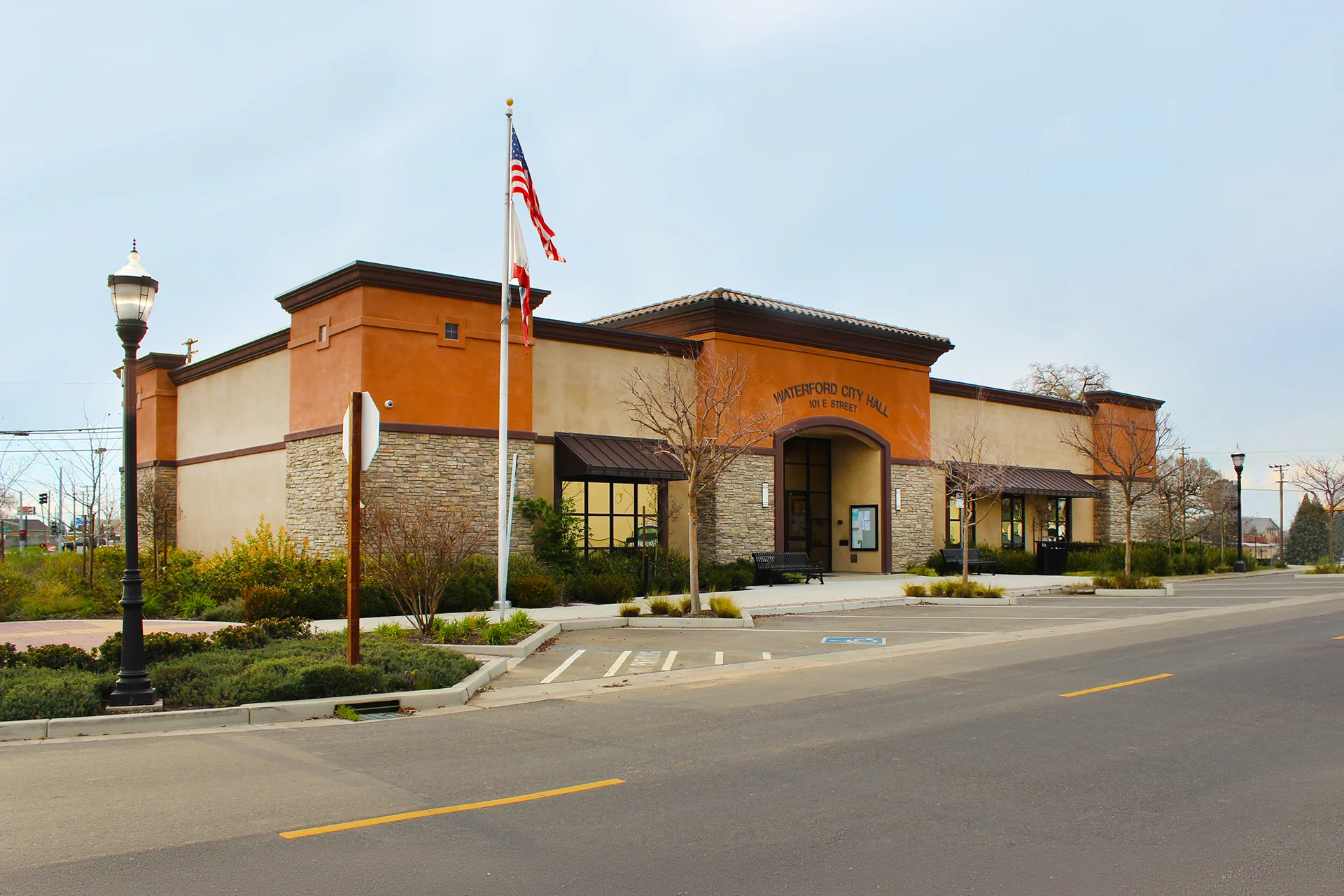 Angle view of the completed Waterford City Hall structure under a cloudy day sky, displaying the arched main entryway, asphalt parking lot lines, deep brown metal window awnings, and an ornamental black metal street lamp post.
