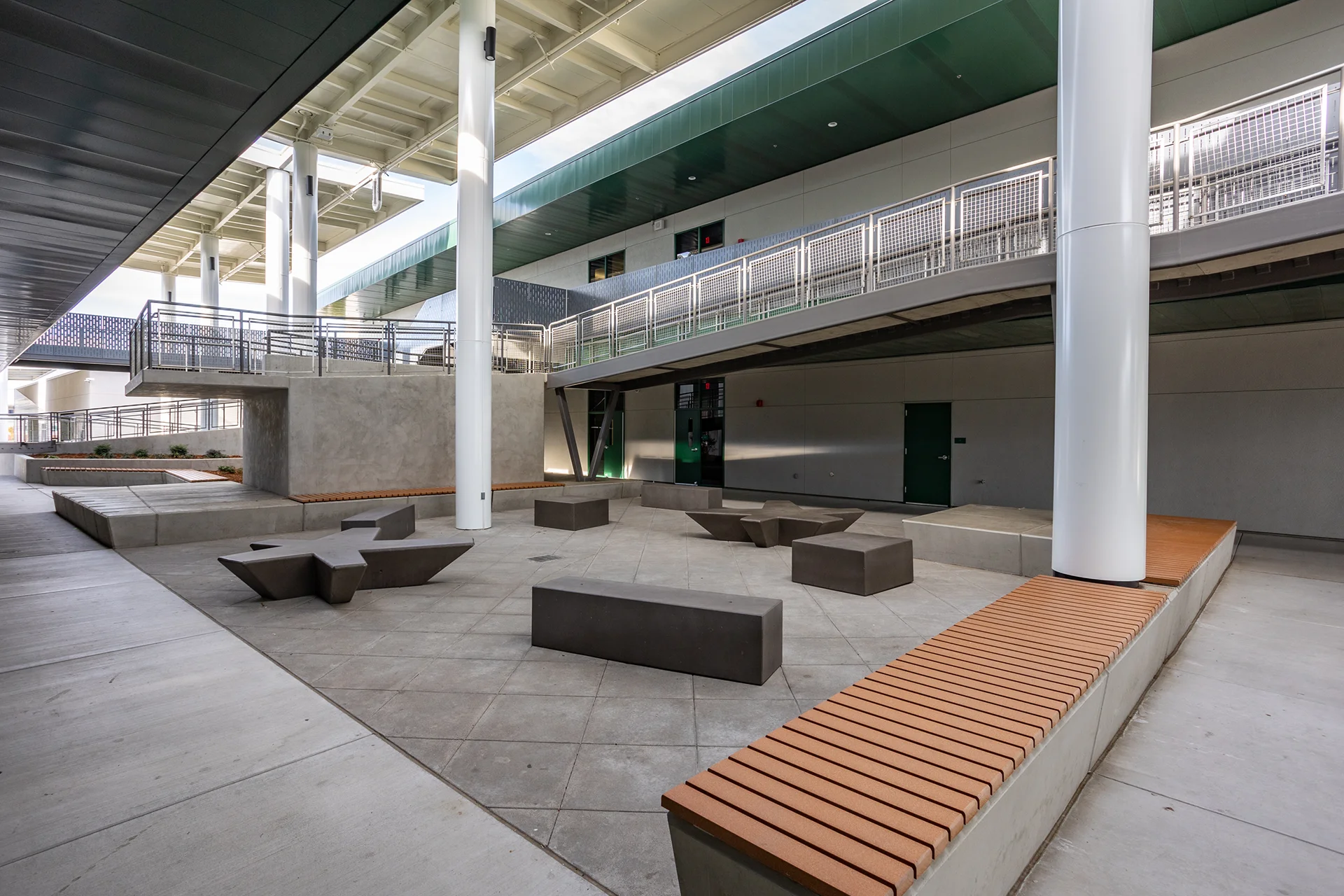 Open-air campus common area in Manteca, California, highlighting geometric concrete paving, architectural star-shaped and rectangular cast concrete benches, wood-slat planter seating, and two stories of covered academic walkways supported by large white structural columns.