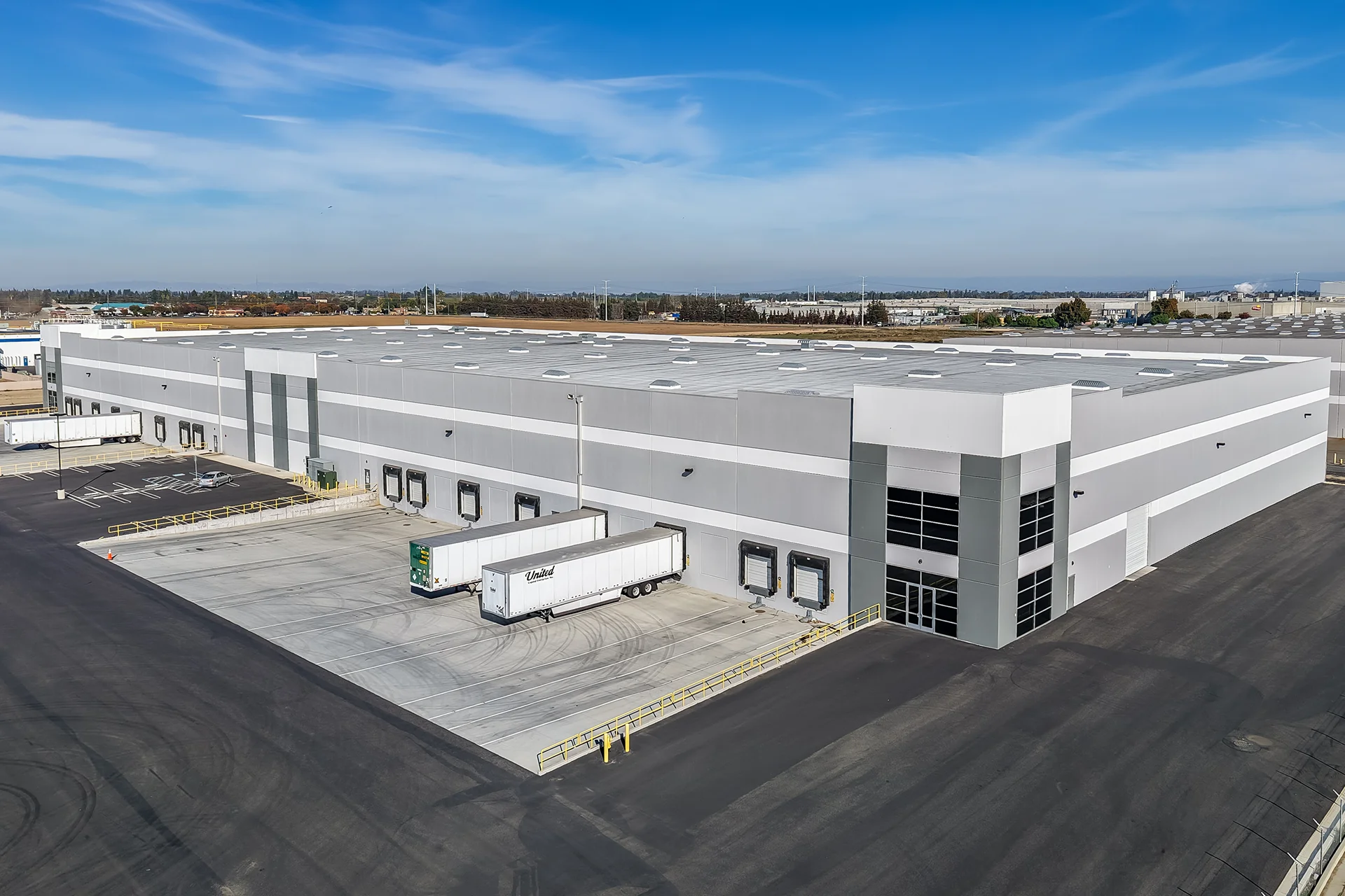 Three-quarter aerial shot highlighting the corporate office corner entryway of a large logistics facility, featuring multi-tier curtain wall window glazing with dark aluminum mullions, structured architectural accent canopies, and logistics trailers backed into active shipping bays.