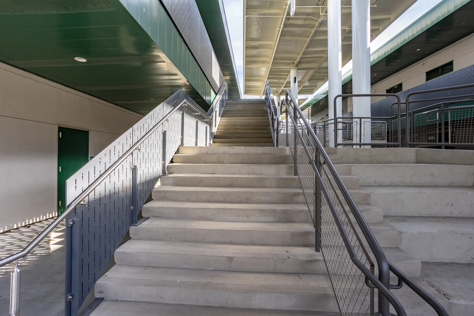 Low-angle view looking up a dual-flight exterior concrete staircase on a modern school campus, featuring brushed stainless steel handrails, custom perforated metal safety guardrails on the lower landing, and woven wire mesh railings on the upper level under a metal canopy.