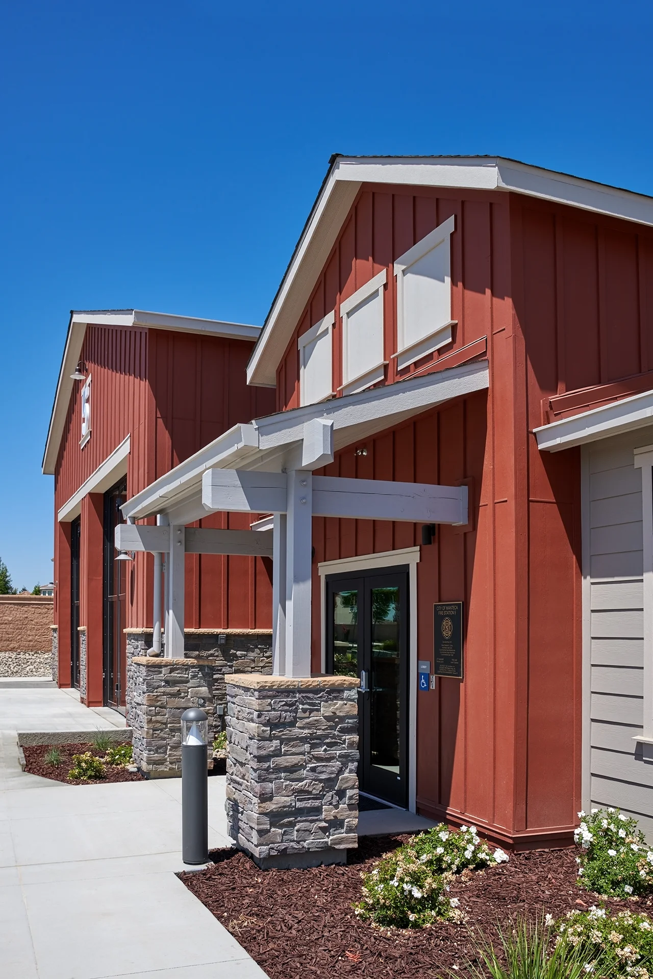 Close-up of the municipal building exterior entrance showcasing a gray wood structural canopy trellis, thick square stone veneer pillar bases, vertical red siding, and dark metal storefront framing around the entrance doors.