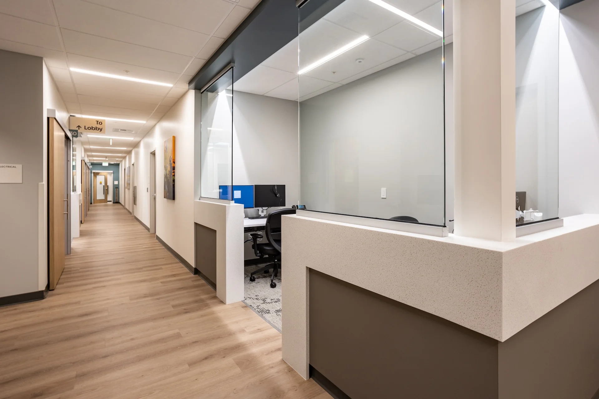 Looking down a smooth healthcare facility corridor featuring premium wood-look vinyl plank flooring, integrated structural privacy glass screens framed by durable solid-surface check-in counters, and low-voltage LED linear ceiling slot lights illuminating guidance signs.