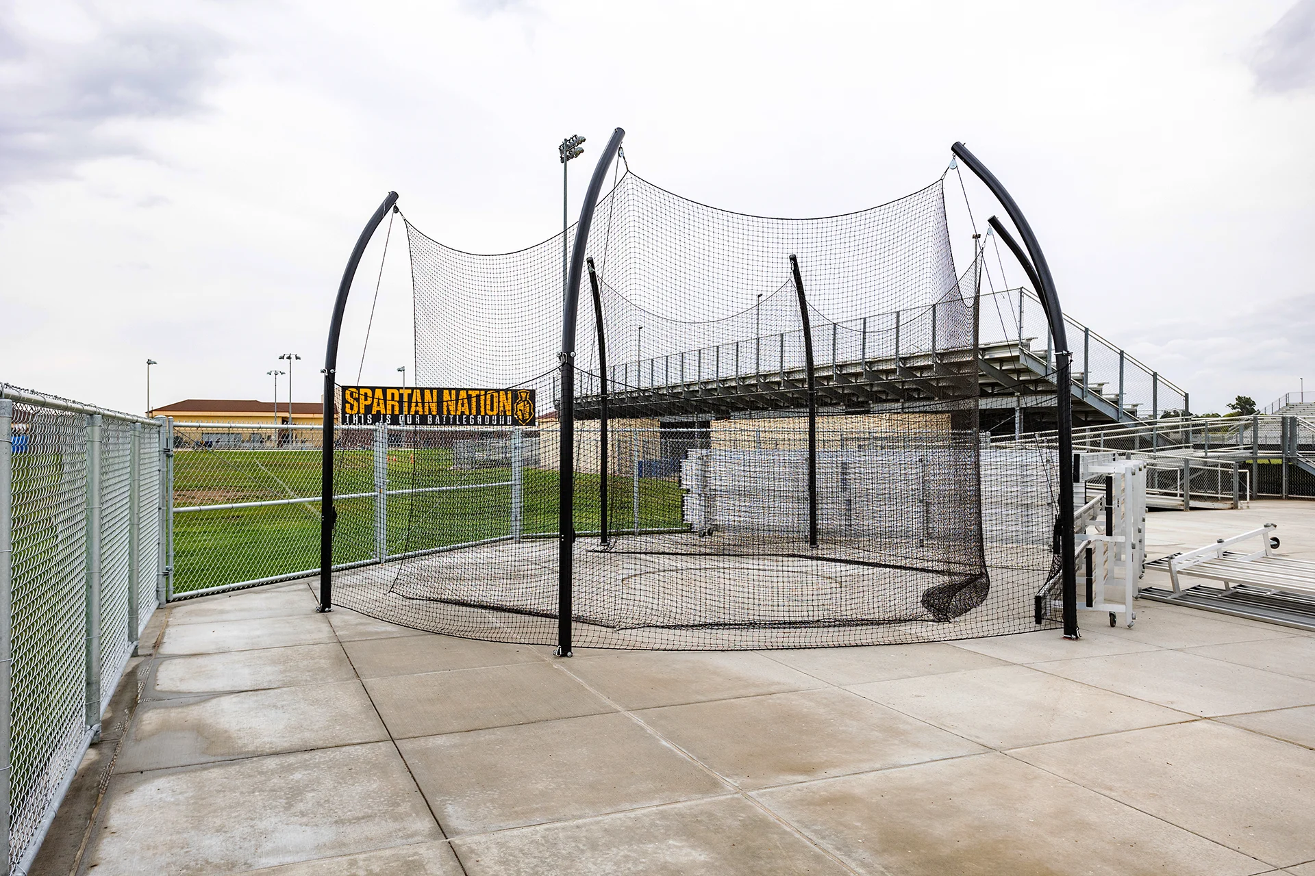 Wide view of a professional athletic discus throwing cage with black netting and support posts next to stadium bleachers.