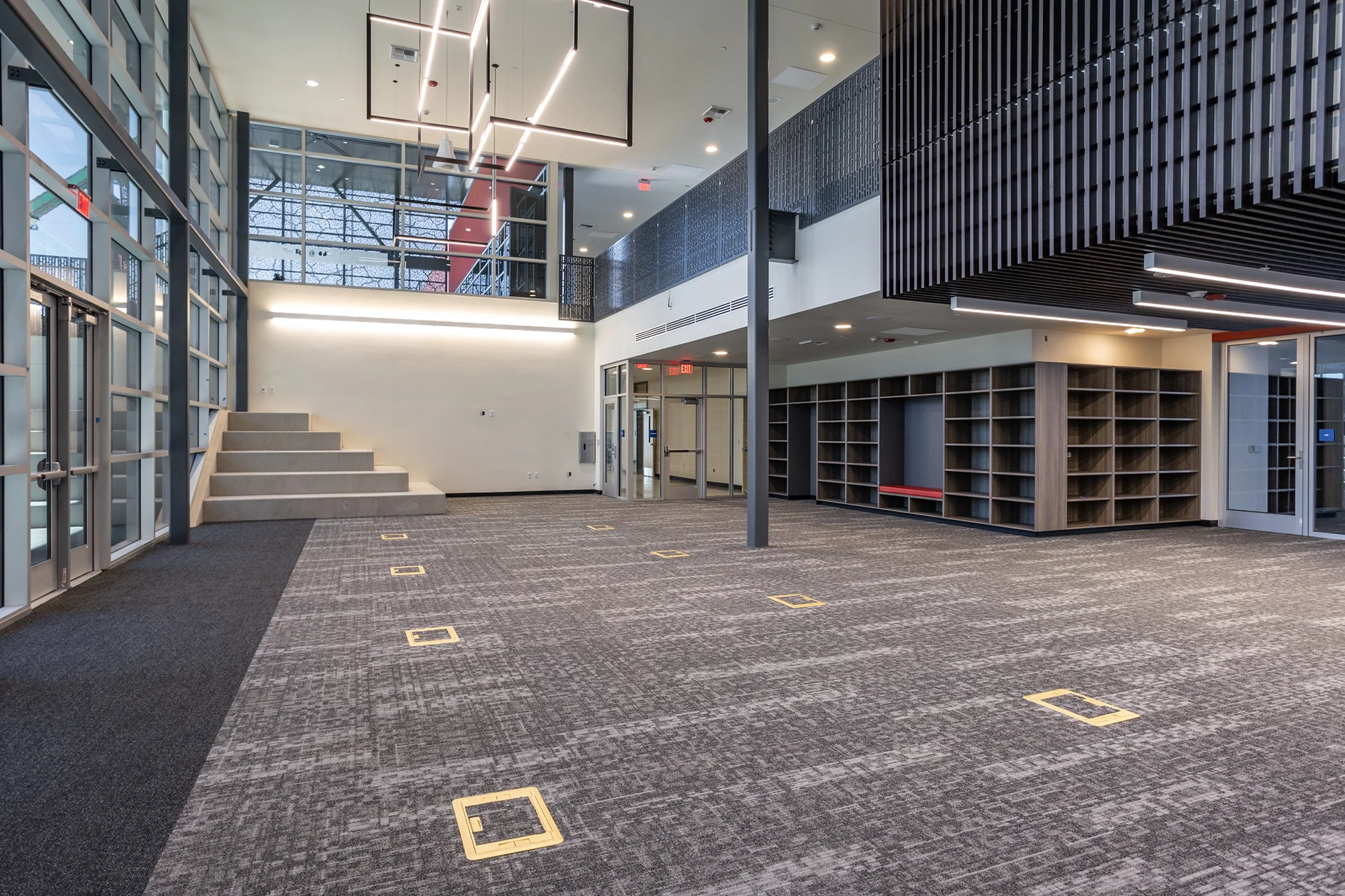 Modern multi-level school atrium featuring a double-height ceiling with custom geometric pendant lighting fixtures, architectural steel support columns, an integrated concrete presentation staircase, built-in wooden locker nooks, and gray commercial carpet tiles.