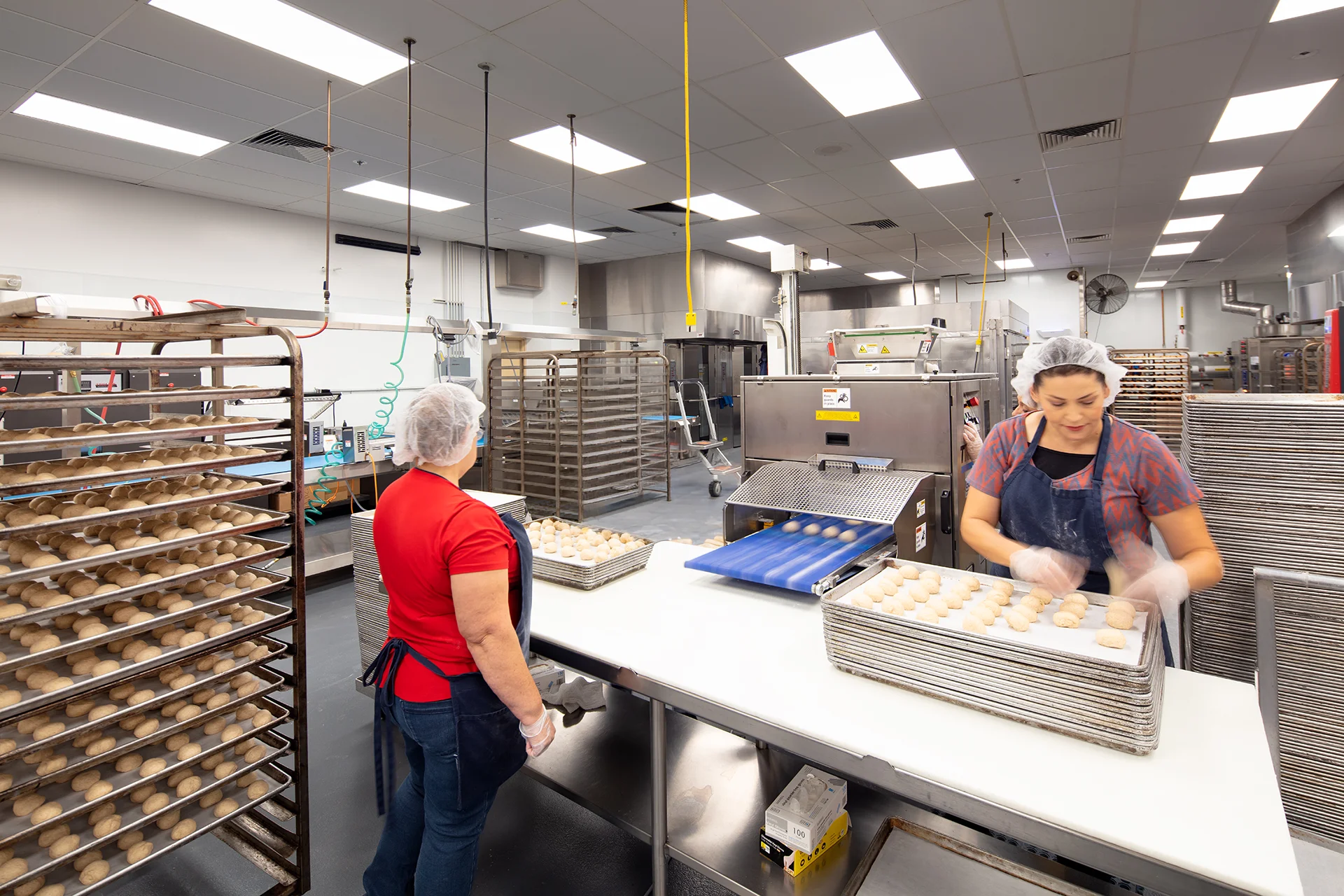 Commercial bakery processing area featuring staff managing an automated dough ball portioning machine, a blue conveyor belt system, and mobile aluminum bun racks under bright recessed LED ceiling panels.