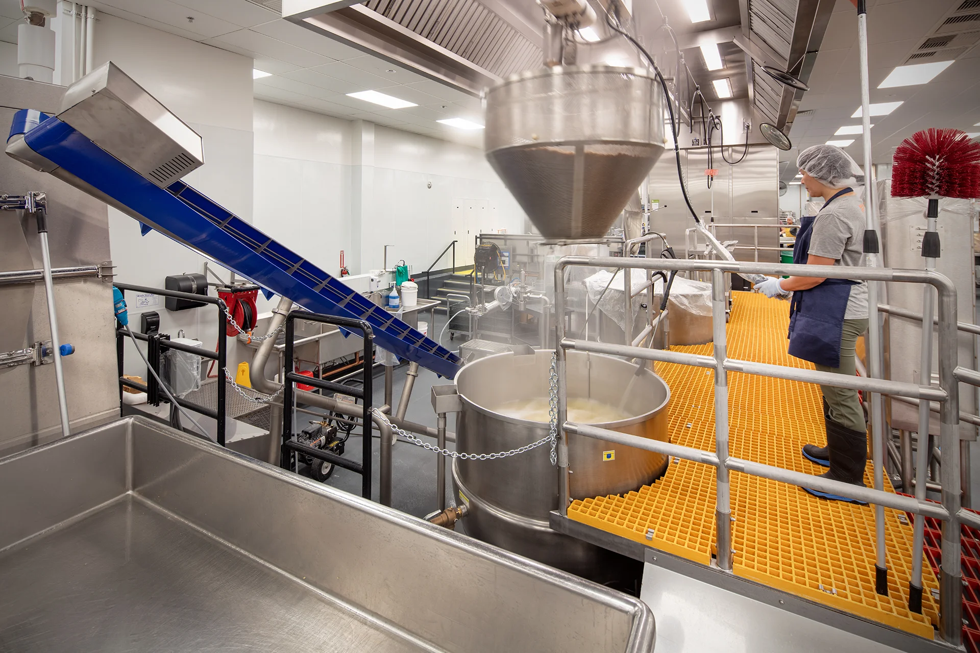 Institutional food preparation facility showing an operator monitoring an automated processing line with a stainless steel steam kettle, overhead ventilation system, and a blue inclined conveyor belt feeding ingredients.