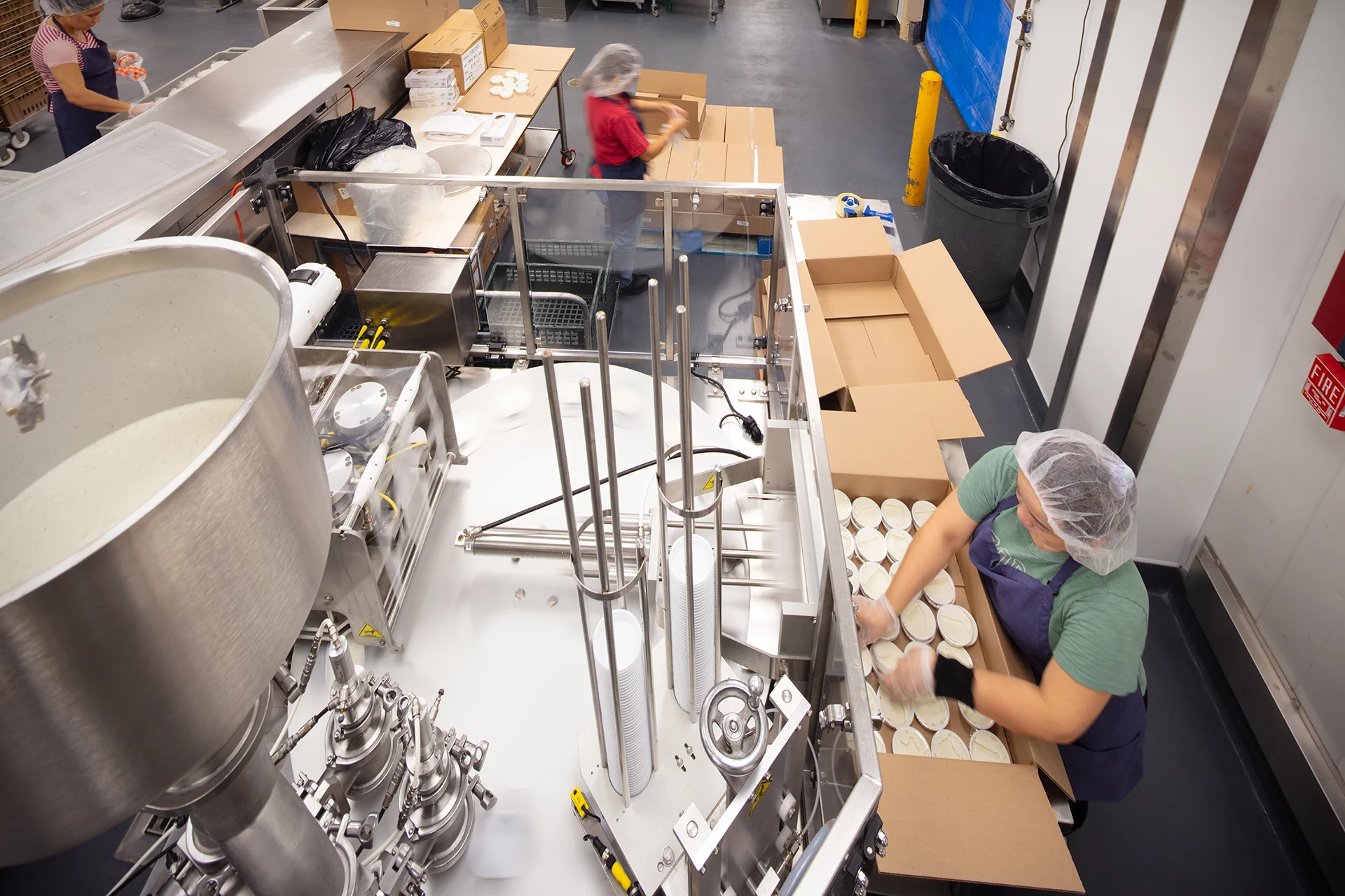 Elevated view of an industrial rotary packaging machine filling and sealing small food containers, with an assembly worker boxing completed items for distribution in an Elk Grove facility.