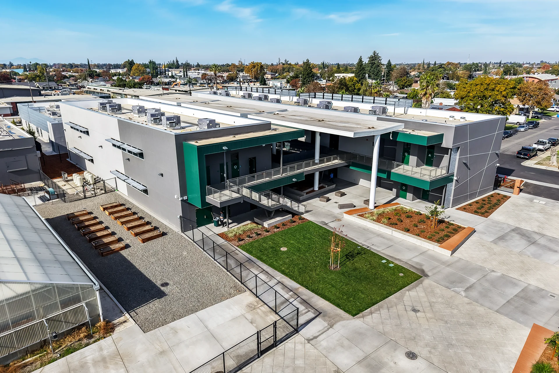 High-angle aerial perspective looking down at a modern educational campus courtyard, displaying square concrete pavers, a square green grass lawn with a newly planted tree, built-in concrete bench planters with wood seating, an agricultural garden area with wood grow frames, and a glass greenhouse building.