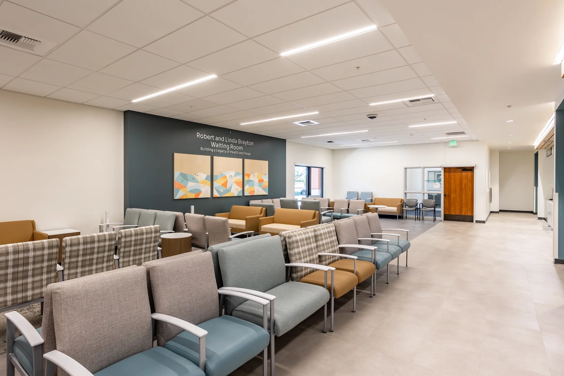 Wide-angle interior shot of a spacious healthcare facility waiting room showing rows of modular medical-grade armchairs in gray, blue, and plaid patterns, an accent wall adorned with a three-panel geometric art installation, linear LED slot light fixtures, and a natural wood door leading to patient corridors.