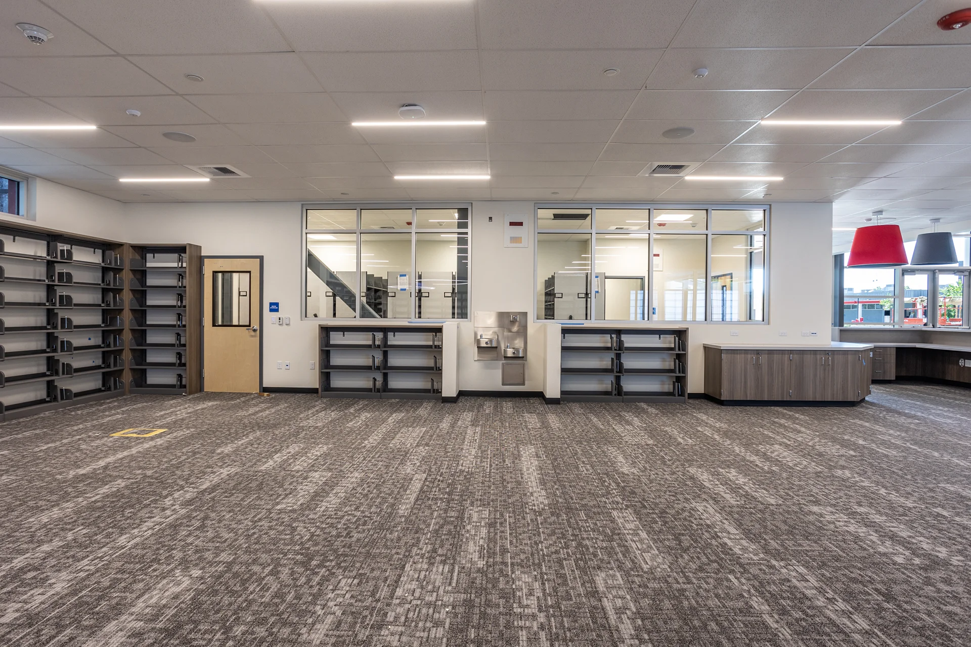 Commercial high school media center renovation in Manteca, California featuring built-in wooden bookshelves, integrated electrical outlets, low-profile linear LED ceiling lights, gray patterned carpet tile, and an indoor drinking fountain station.