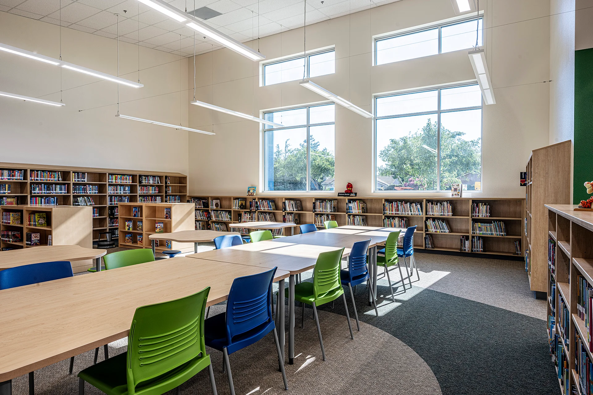 Modern elementary school library interior constructed by CT Brayson & Sons, featuring large multi-pane windows for natural lighting, light wood library bookshelves, long study tables paired with vibrant green and blue plastic chairs, linear LED suspended light fixtures, and multi-toned commercial carpeting.