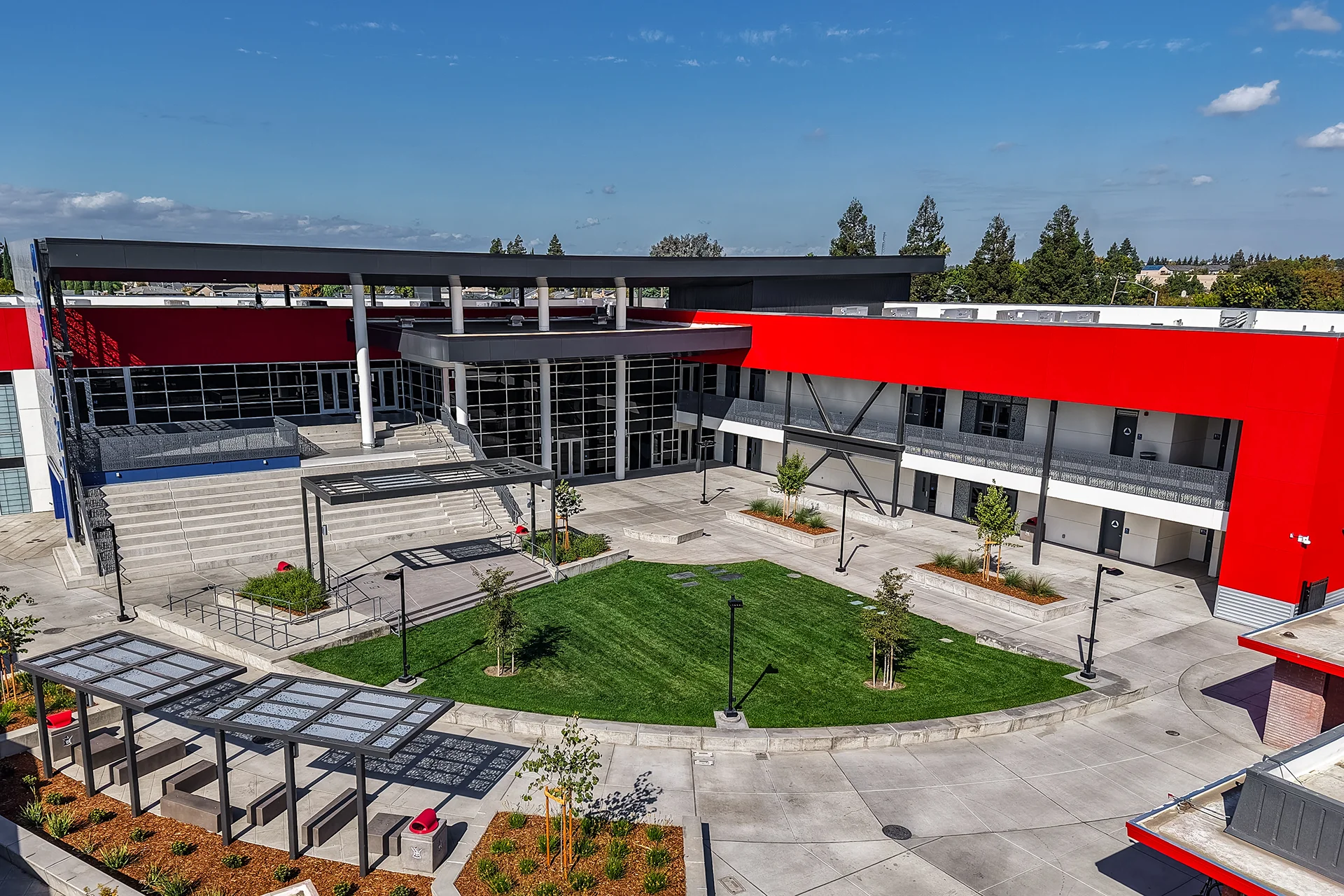 Institutional high school courtyard development in Manteca, California featuring a multi-story glass storefront curtain wall, industrial steel framing accents, a central green quad lawn, and matching modern pedestrian site amenities.