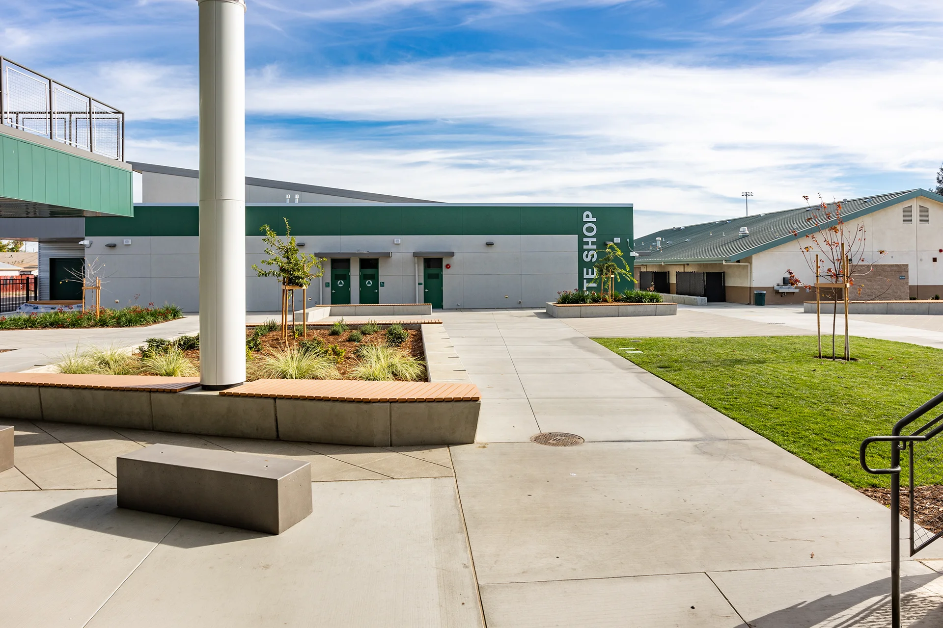 Looking across a modern school campus plaza in San Joaquin County, featuring geometric concrete paving, built-in cast-in-place concrete planter benches with wood slat tops, decorative native grass landscaping, and the dark green exterior of a vocational site shop facility.