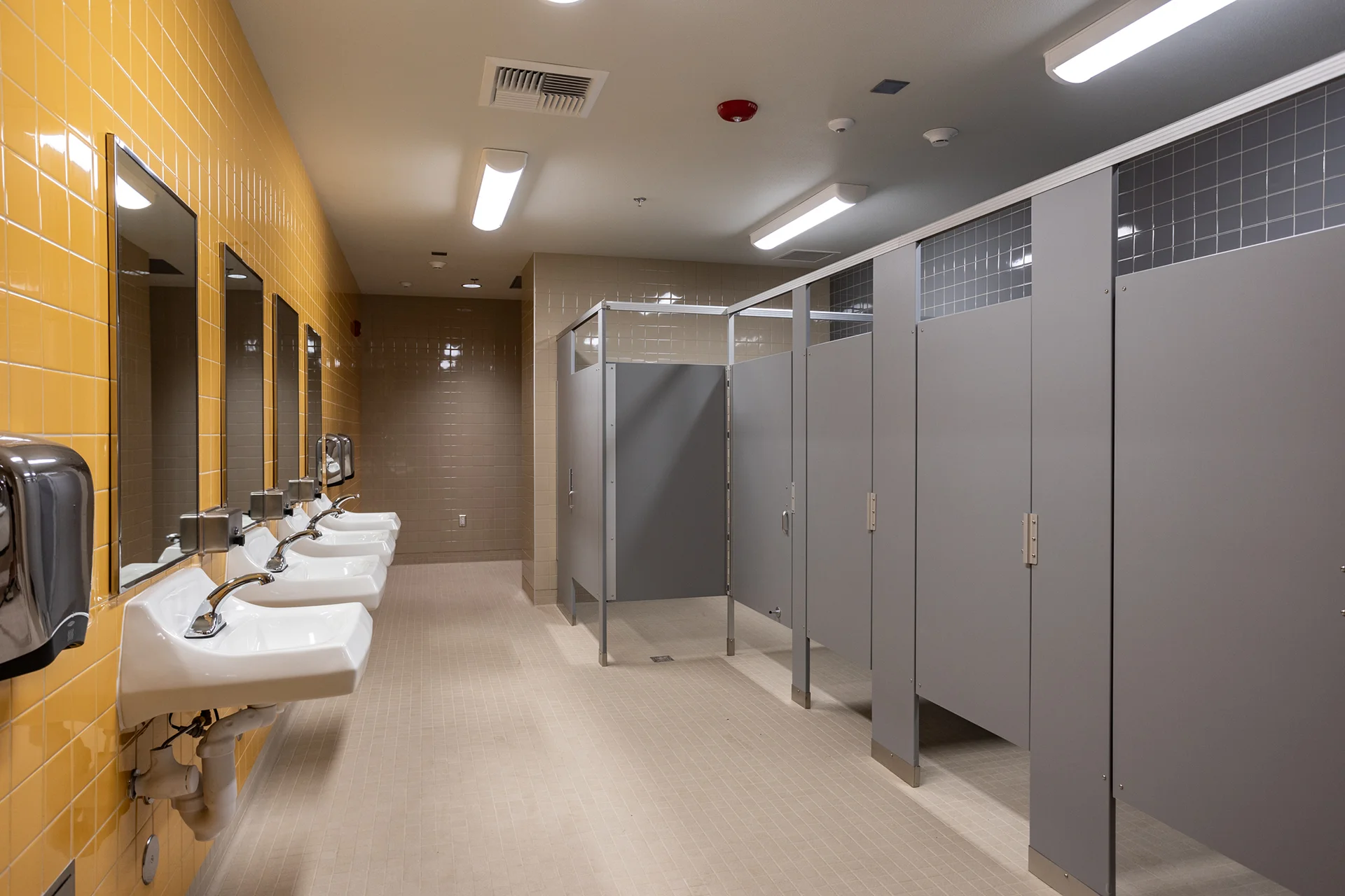 Commercial public restroom facility built with a long wall-hung array of white porcelain washbasins equipped with chrome automatic sensor faucets. The space features vibrant yellow gloss ceramic wall tiles, rectangular vanity mirrors, and a row of floor-anchored grey plastic laminate toilet partition stalls.