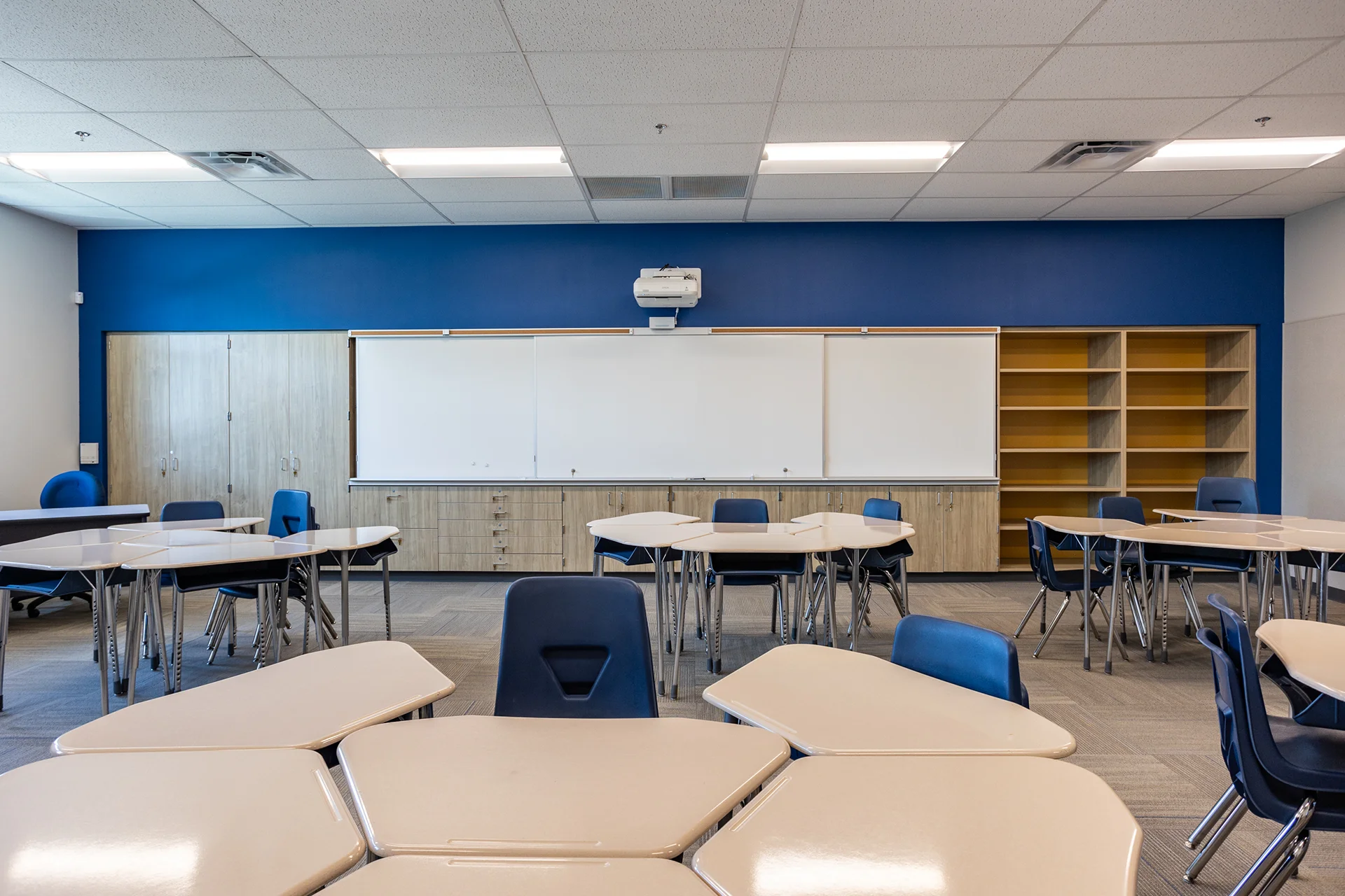 Front perspective of an institutional classroom displaying a long white board system integrated into custom light oak cabinetry drawers and closets. The room is finished with gray carpet tiles, ergonomic blue student chairs, matching geometric desks, and a royal blue focal wall under bright troffer ceiling lights.