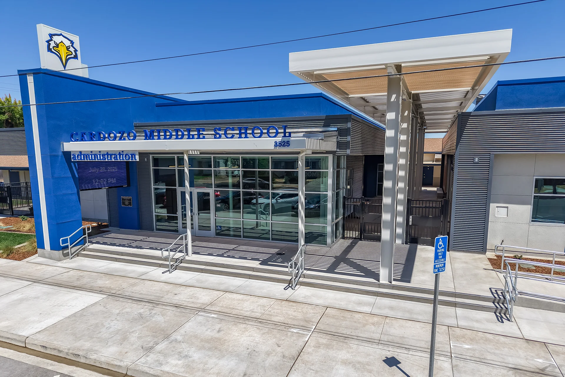 Elevated view of the modern administrative entrance showing concrete approach pathways, a digital LED directory or announcement display screen integrated into a blue architectural column, steel security gates leading into an interior corridor, and a blue metal ADA handicap parking sign post.