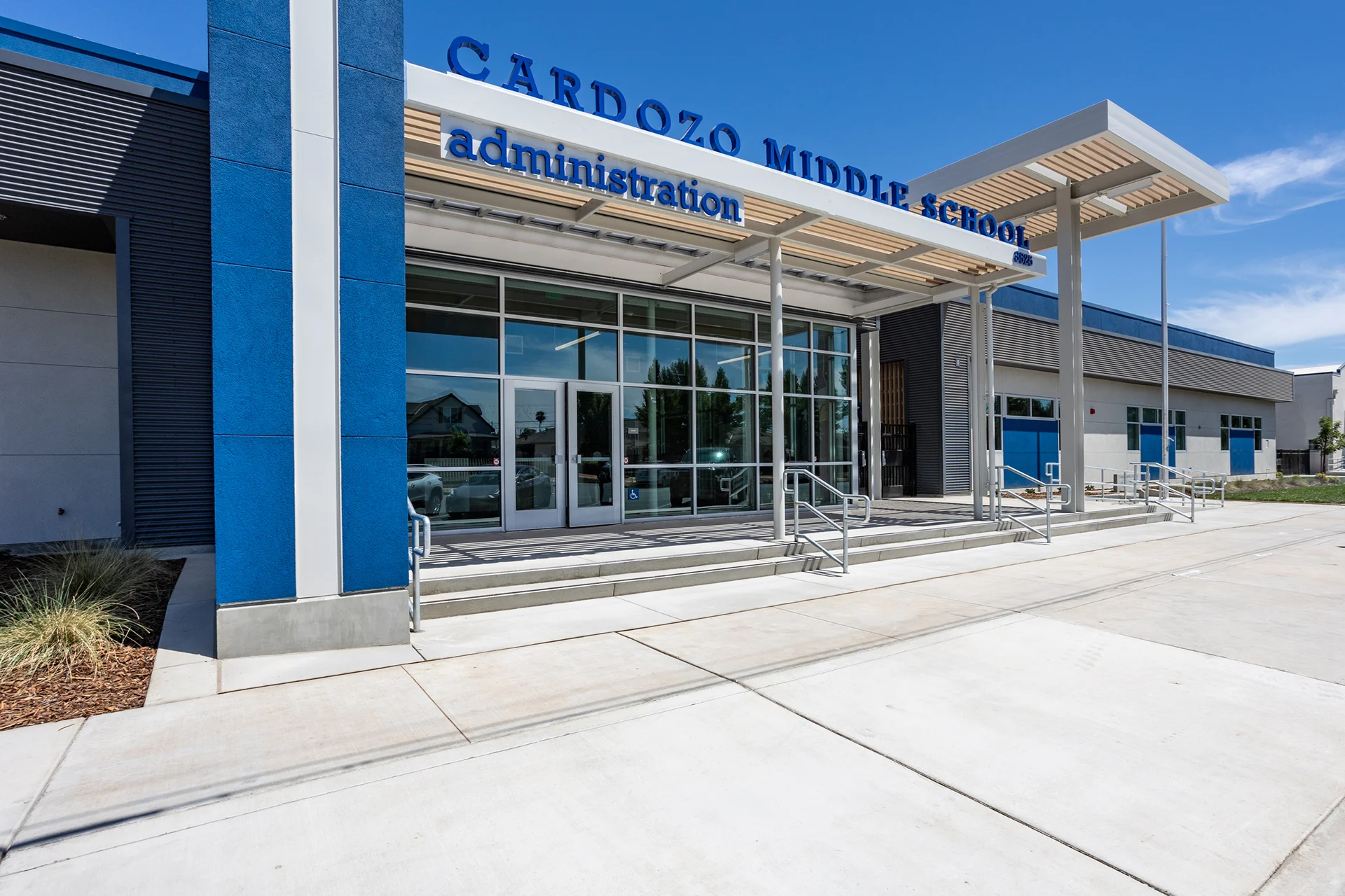 External perspective of the Cardozo Middle School administration building entrance, showcasing a contemporary structural design by CT Brayson & Sons. The facade features deep blue stucco pillars, aluminum curtain wall glass storefront windows, dimensional exterior building signage, and an extended steel canopy awning with light wood-paneled slats over concrete steps and an ADA-compliant handrail system.