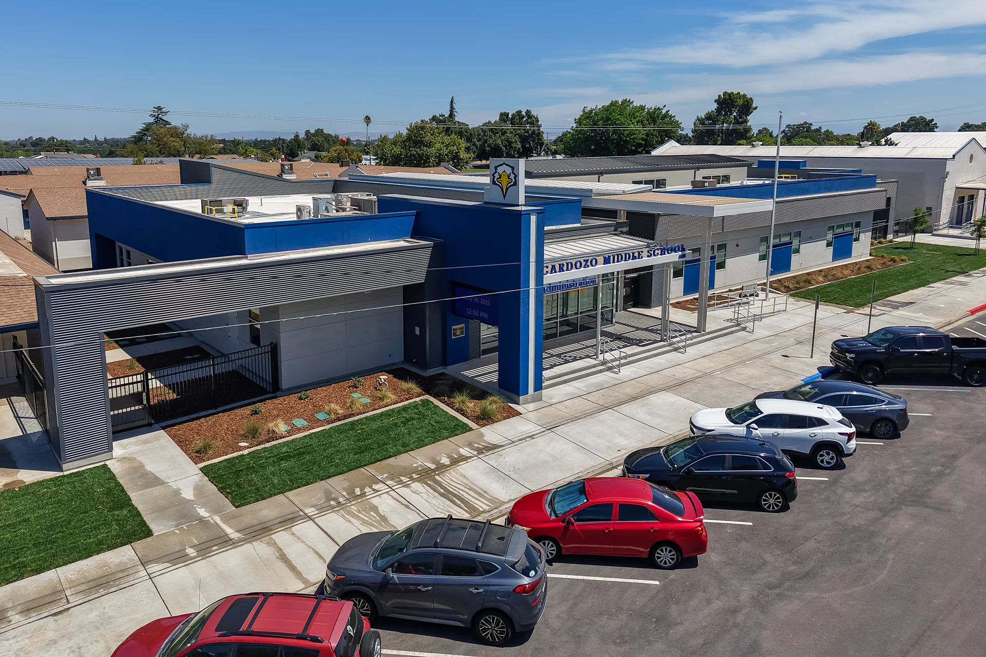 High-angle drone aerial view of the completed Cardozo Middle School administrative complex in Riverbank, California. The image captures the modern blue-and-grey architectural contours, a low-profile flat roof with industrial HVAC installations, a prominent monument sign base featuring the eagle mascot, a concrete entry plaza, and an adjacent striped asphalt staff parking lot.