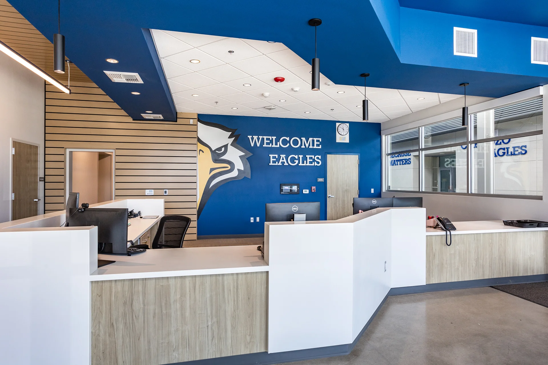 Interior of the Cardozo Middle School main administration lobby, constructed by CT Brayson & Sons. The space features a custom angular reception desk with a wood-grain and white solid-surface finish, a vibrant blue accent wall reading Welcome Eagles alongside a large eagle mascot graphic, suspended black cylindrical pendant lights, and an architectural geometric drywall ceiling design.