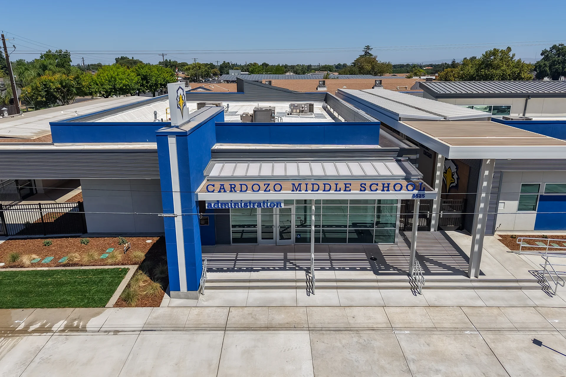 Aerial shot focusing on the architectural entrance of the administration suite, showcasing a commercial storefront glass entryway system beneath a protective corrugated metal roof canopy with structural column supports, balanced by off-white stucco panels and an integrated blue accent tower.