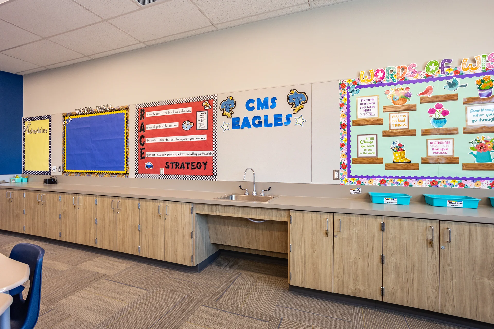 Close-up interior view of a middle school classroom utility center, detailing long laminate storage cabinets and a drop-in stainless steel sink with a gooseneck faucet. The counter installation includes an open wheelchair knee-clearance cutout for ADA compliance below active student words of wisdom bulletin boards.