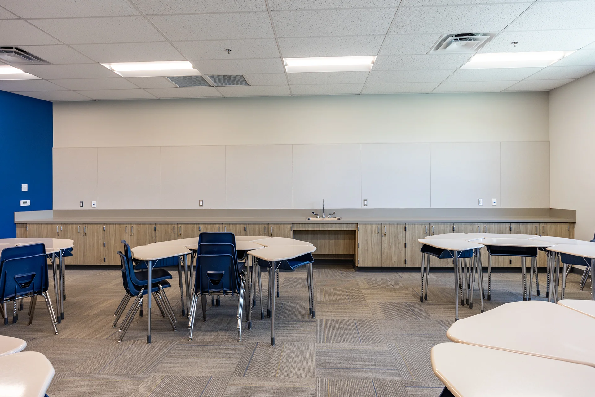 Academic science or resource room layout showcasing a continuous wall-to-wall run of built-in laminate cabinets and utility counters featuring an integrated steel sink station. The classroom area contains multiple arranged groupings of tan geometric student desks paired with dark blue plastic shell chairs.