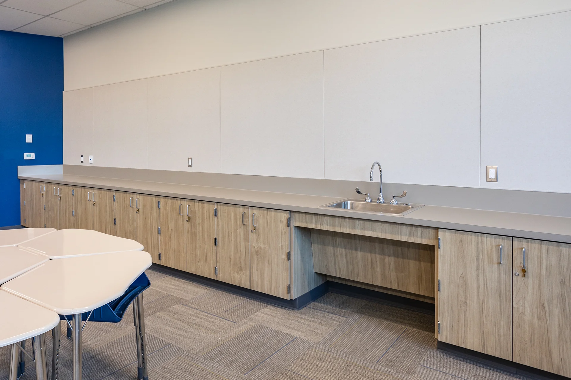 Institutional classroom wet bar station built with an extended line of wood-grain laminate lower cabinets and a smooth grey solid surface countertop. The counter integrates a drop-in stainless steel sink with a gooseneck faucet hardware setup and features an open knee-clearance cutout for wheelchair accessibility.