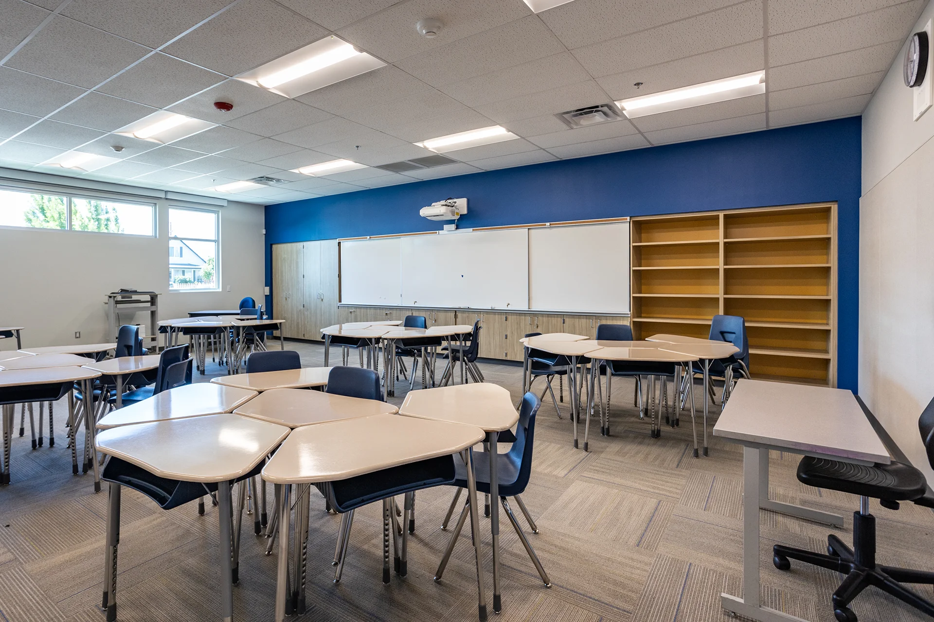 Modern middle school classroom interior featuring modular, wave-shaped student desks grouped for collaborative learning. The educational space is built with an acoustic tile drop ceiling, high-efficiency recessed lighting, a royal blue accent wall, wall-mounted dry erase markerboards with an integrated short-throw projector, and light wood cabinetry.