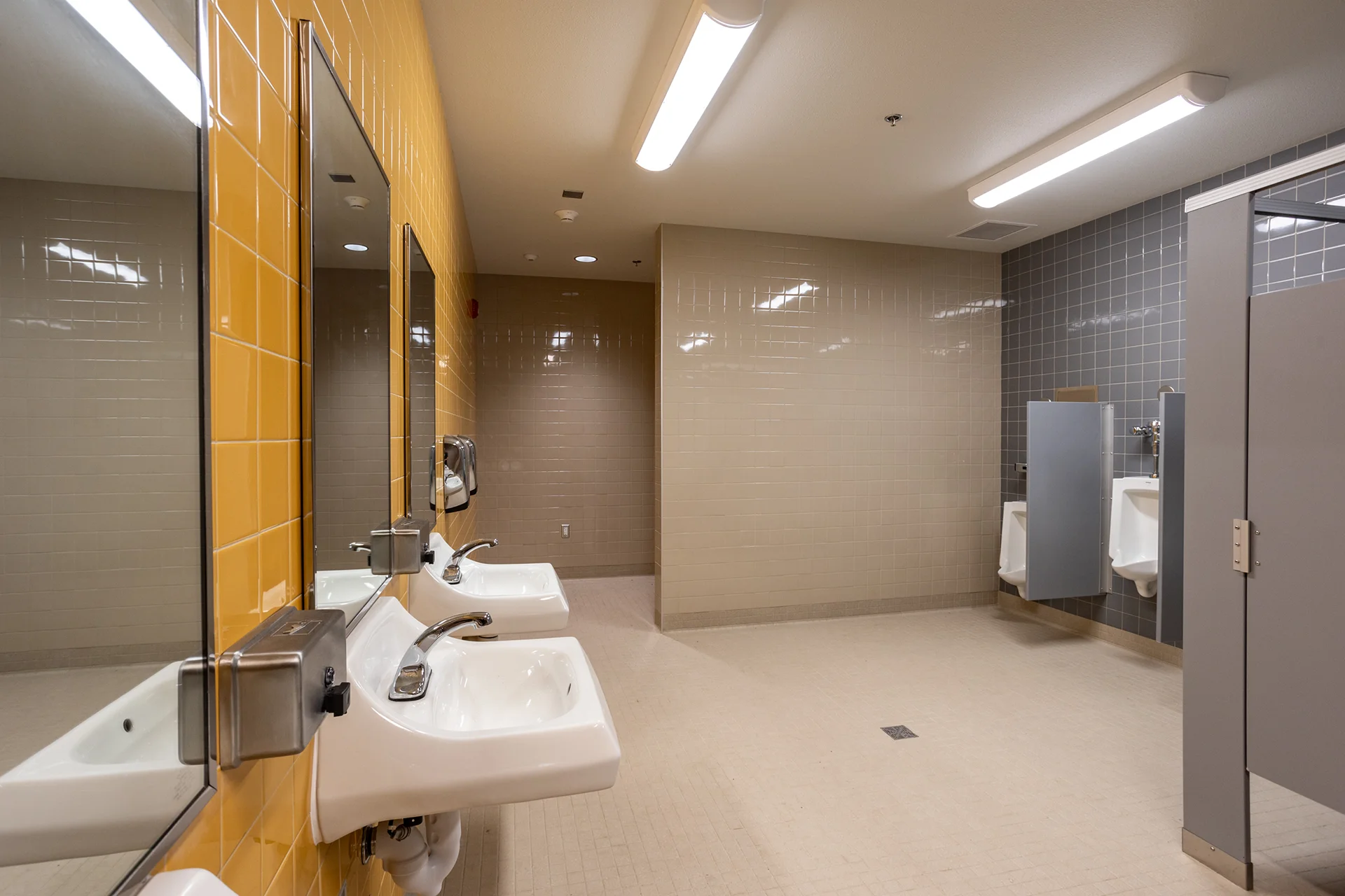 High-traffic school restroom interior showcasing wall-mounted white porcelain sinks fitted with heavy-duty chrome hardware and automatic soap dispensers. The vanity layout features individual frameless rectangular mirrors set against a bright yellow glazed ceramic tile wall facing a neutral tile backdrop.