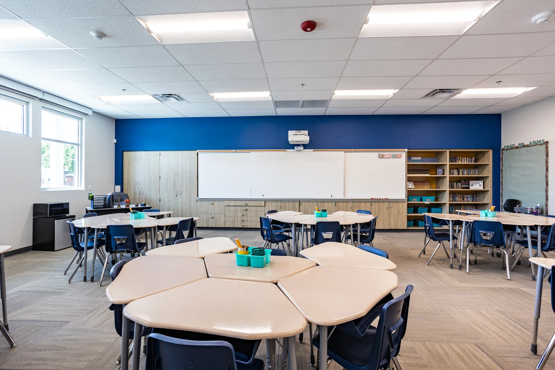 Wide view of a completed collaborative learning classroom, featuring clusters of modular tan desks and matching blue plastic student chairs. The institutional room is finished with grey carpet tile flooring, built-in oak storage millwork, whiteboard tracks, and high-efficiency troffer ceiling lights.