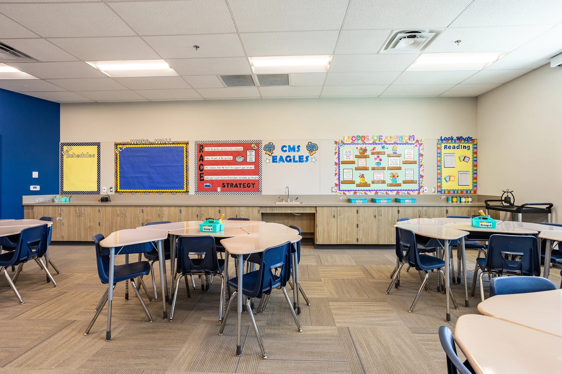 Language arts classroom interior featuring a continuous run of oak base cabinetry topped with a grey laminate utility counter and an integrated steel sink. The wall above is detailed with colorful educational display boards, an ELA word wall, and a custom CMS Eagles logo graphic.
