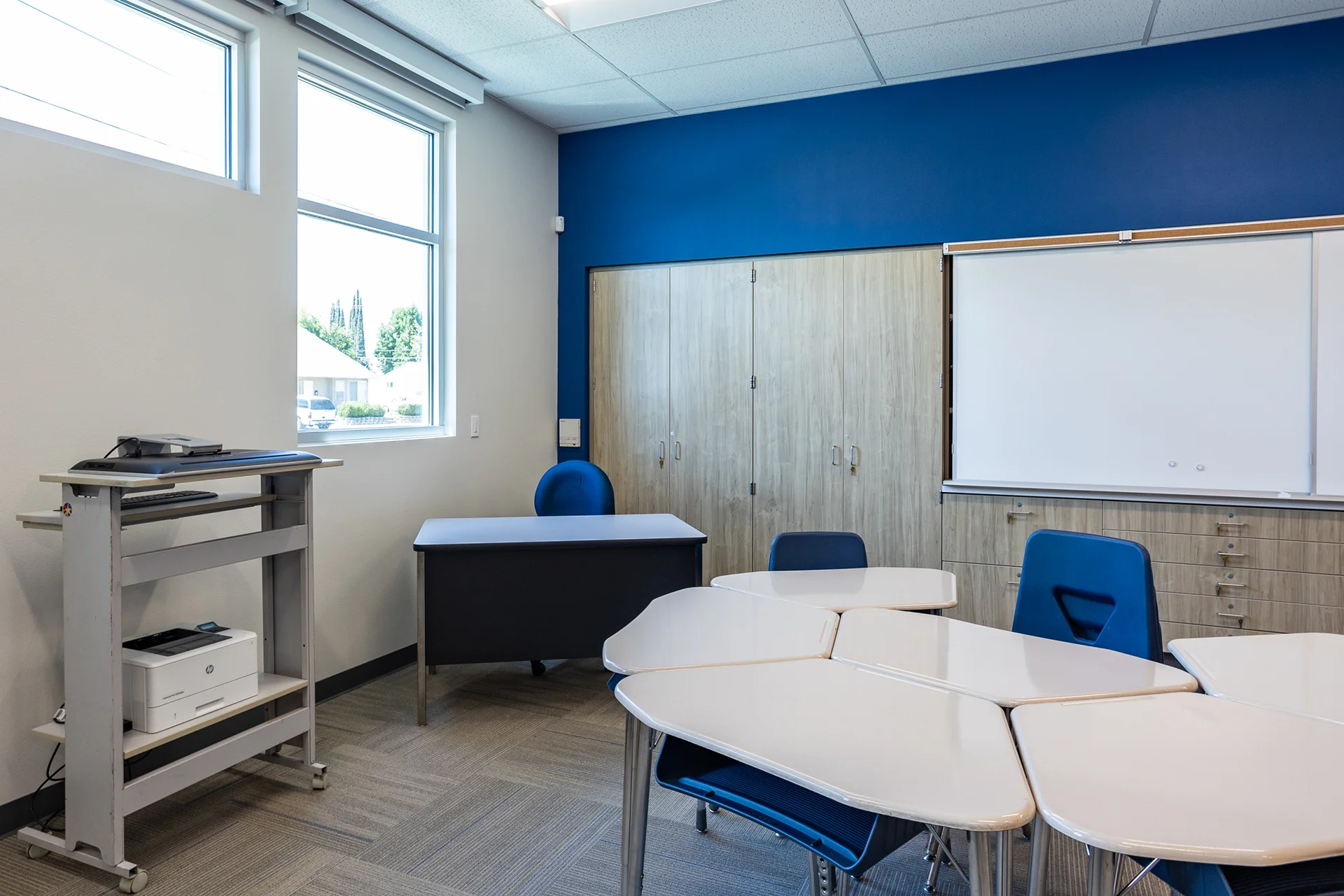 Corner view of an educational classroom showing a dedicated instructor desk paired with an ergonomic blue task chair. The surrounding space highlights a mobile steel rolling utility cart housing a laser printer, storefront glass window units, a large whiteboard array, and built-in vertical storage wardrobes.