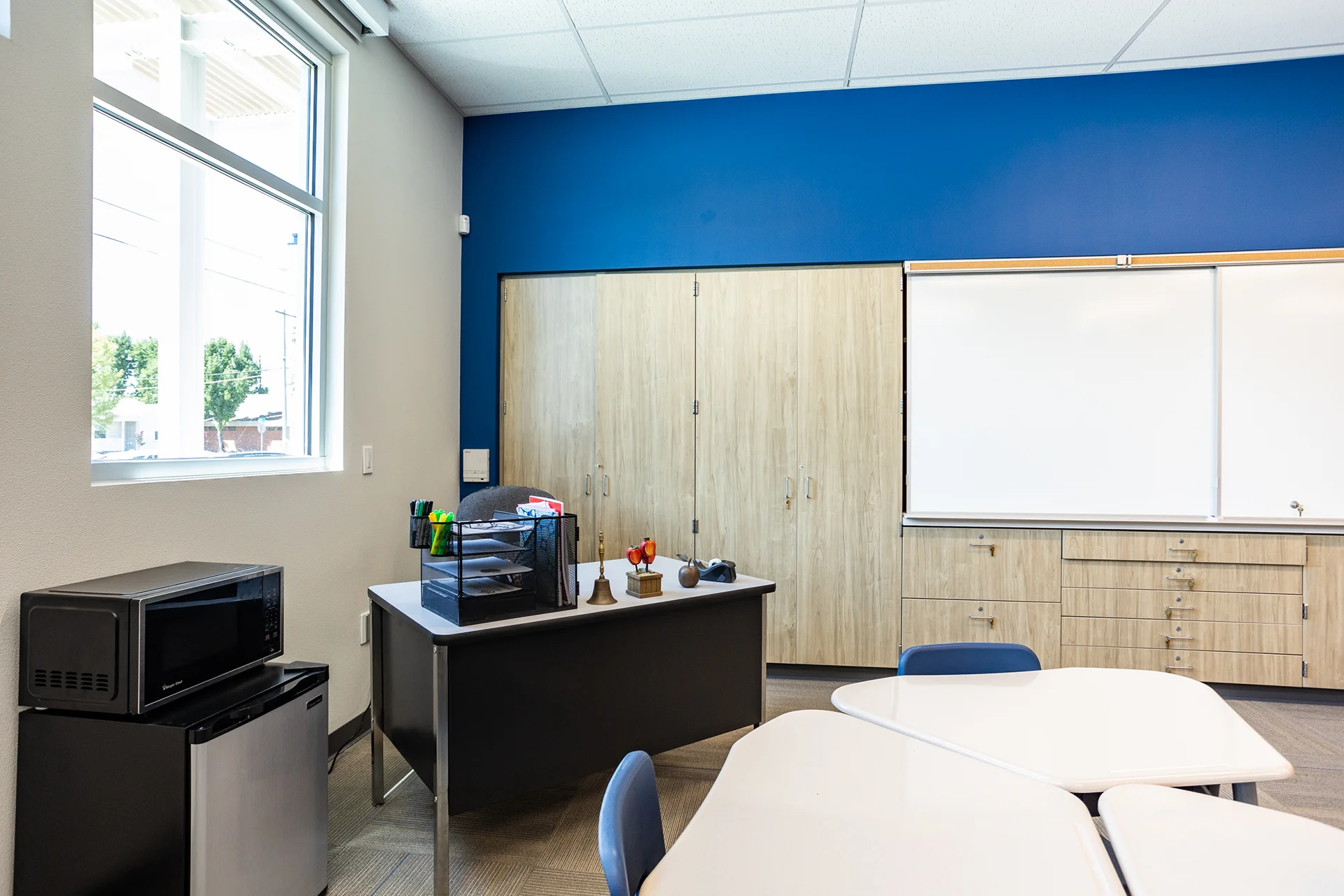 Secondary angle inside an educational classroom highlighting an instructor desk workspace equipped with desktop office organizers, set alongside a compact black microwave and mini-refrigerator appliance station in front of full-height light oak utility closets.