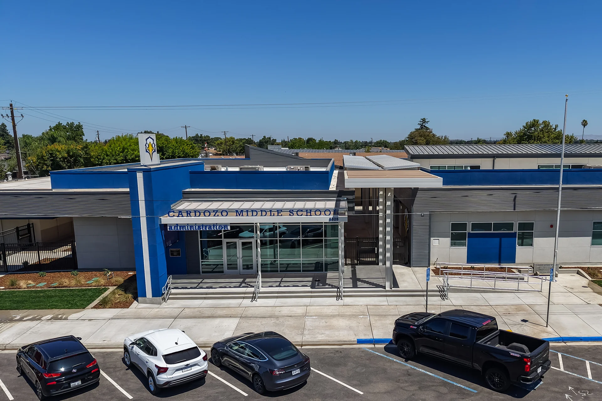 Drone aerial elevation view of the front entrance of Cardozo Middle School. The commercial building design features smooth grey plaster panels, deep blue accent blocks, a large storefront glass window wall under a steel sunshade trellis canopy, structural security gates, and a striped asphalt vehicle parking zone.