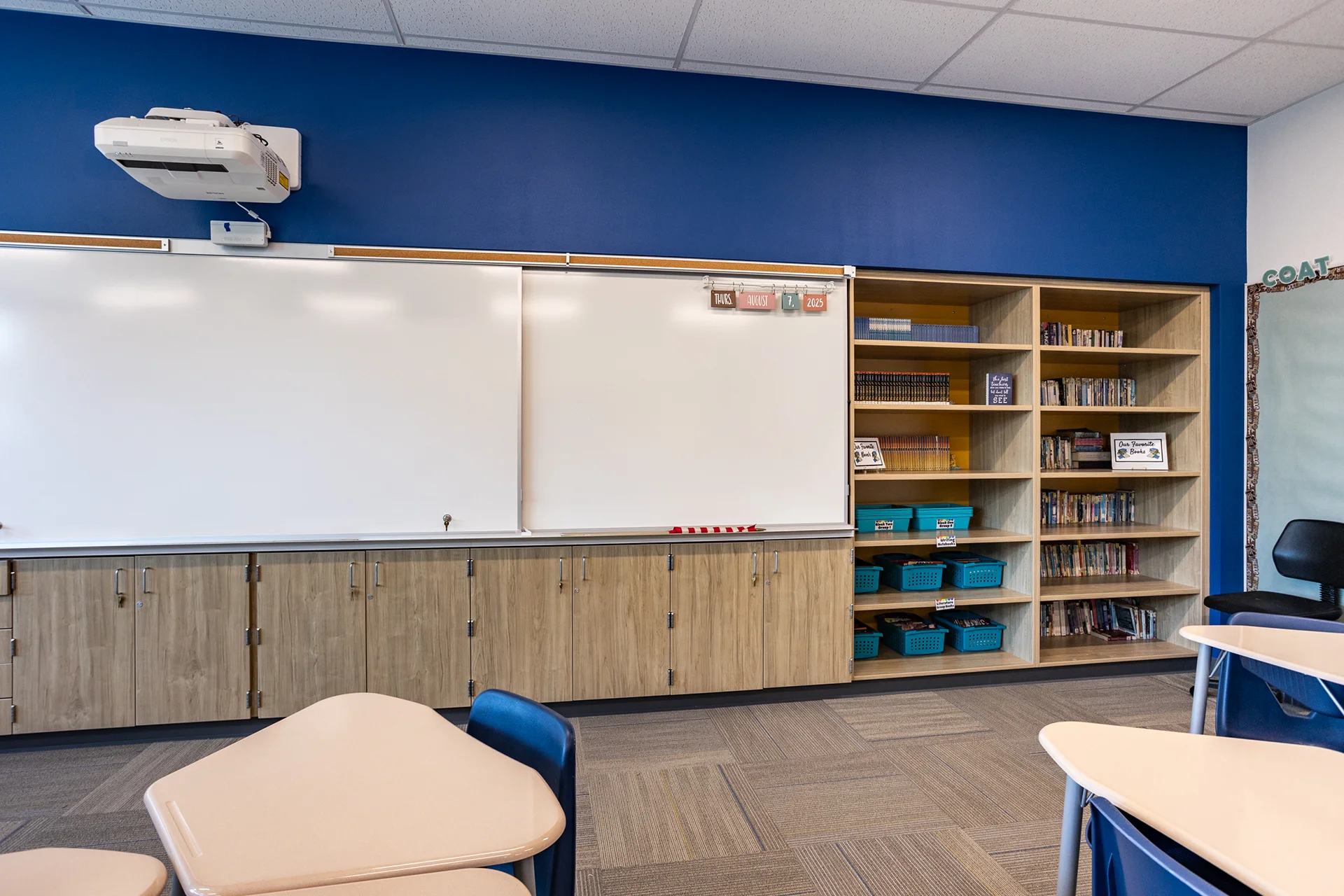 Academic classroom interior showing built-in light wood storage cabinetry and full-height library bookcases stocked with books and sorting bins. The casework sits below an institutional white markerboard, an overhead short-throw media projector, and a cobalt blue focal wall.