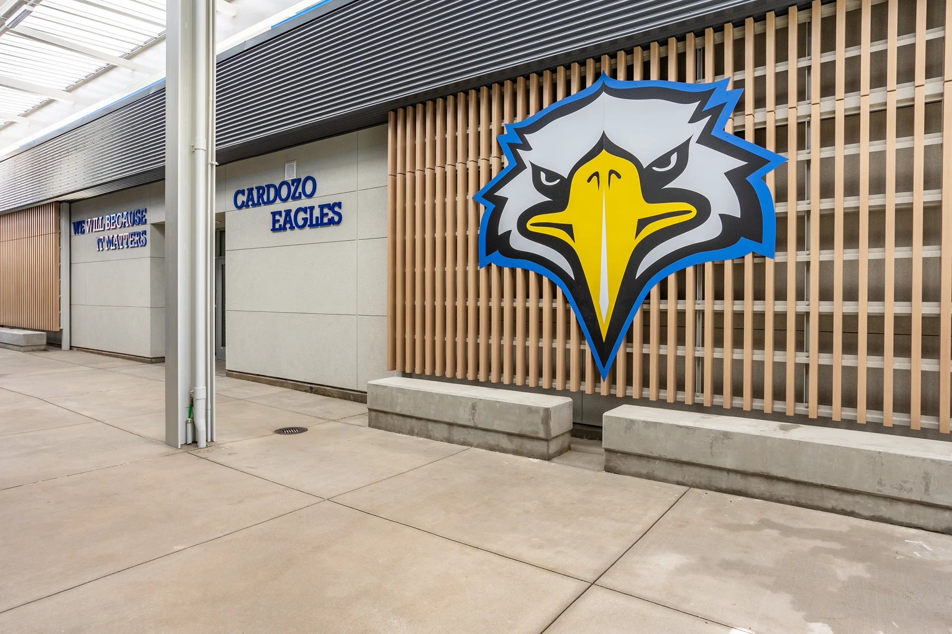Covered campus breezeway courtyard showcasing a large-scale graphic plaque of the Cardozo Eagles mascot logo mounted on a vertical wood slat architectural screen. The school courtyard features smooth concrete flooring, cast concrete bench seats, and distant dimensional building lettering.